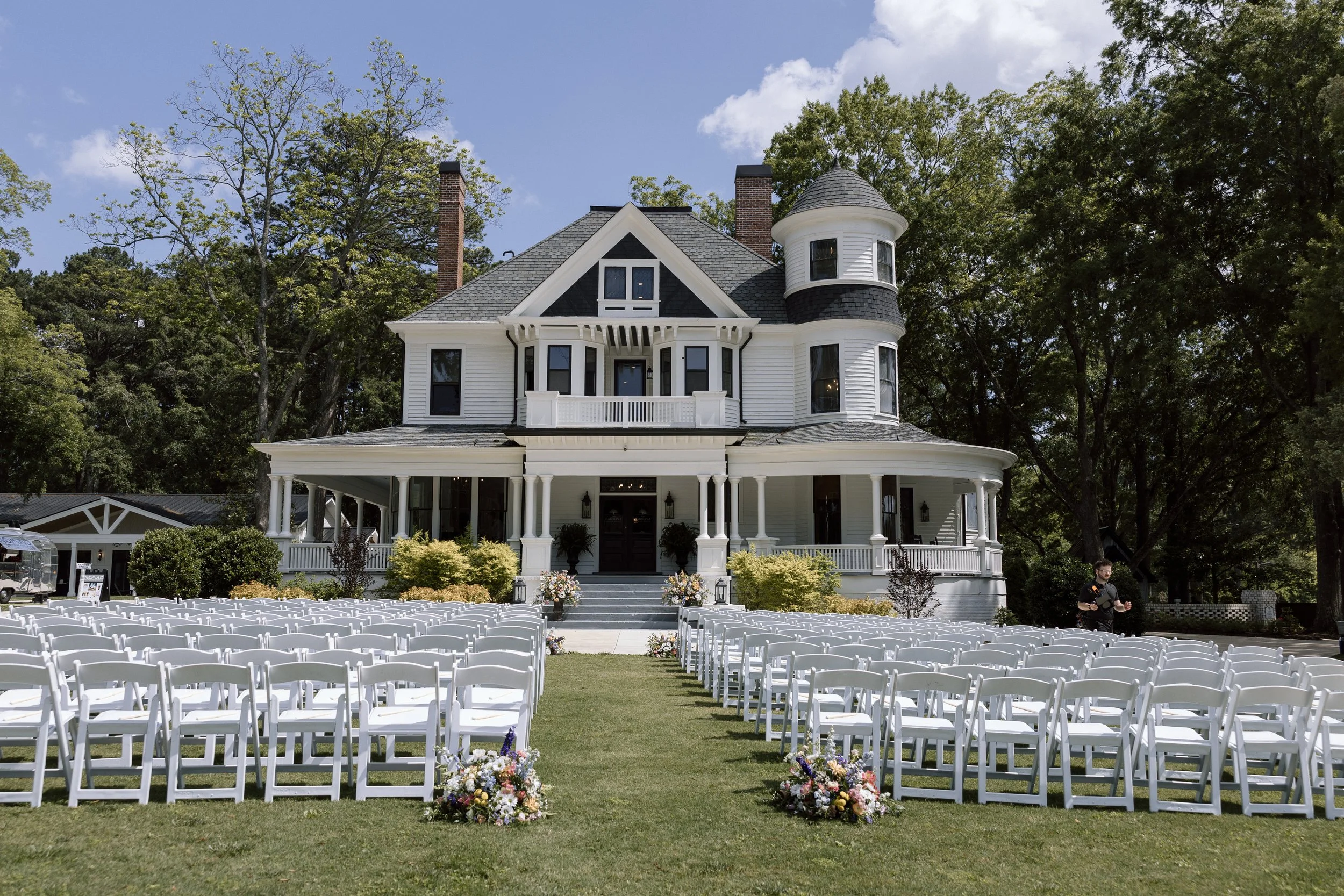 A large white Victorian-style house decorated for a wedding, with white chairs arranged in rows on a lawn and floral arrangements at the front. A man is seen on the right side near the chairs.