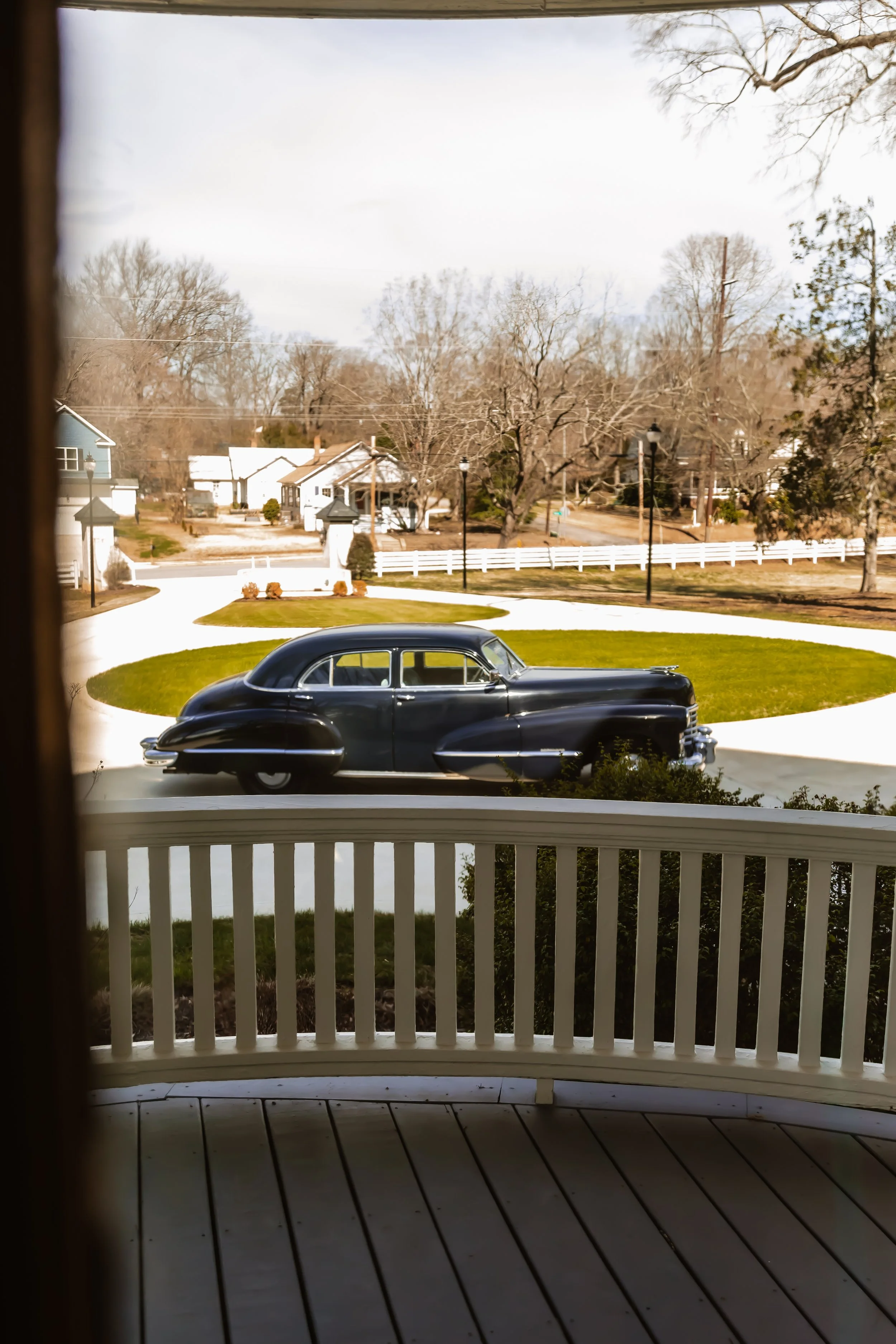 View of a black vintage car parked on a circular driveway in front of a house, seen from a porch with white railings. There are trees and houses in the background on a clear day.