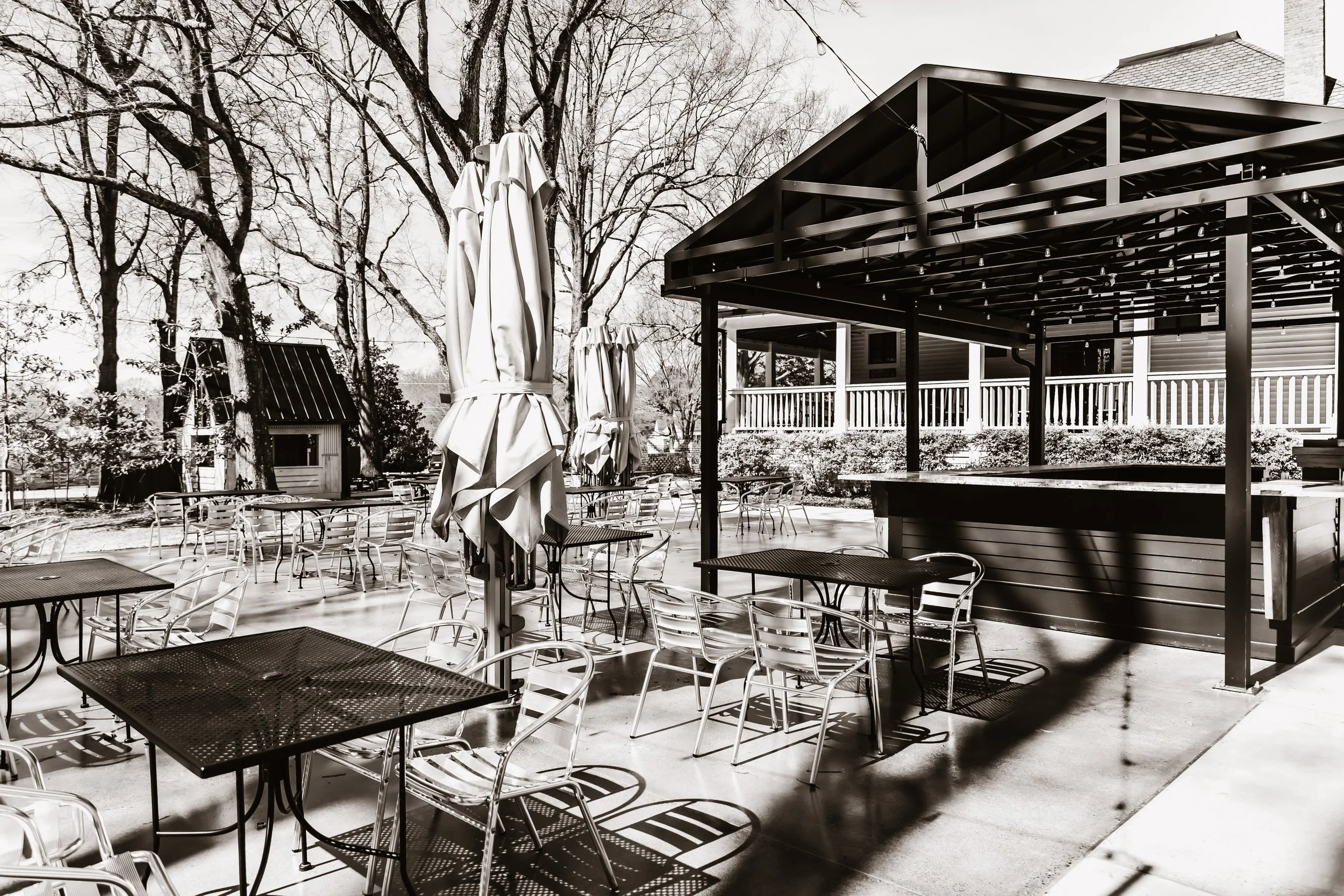 Empty outdoor patio with tables and chairs, umbrellas, a bar area, trees, and a house in the background, in black and white.