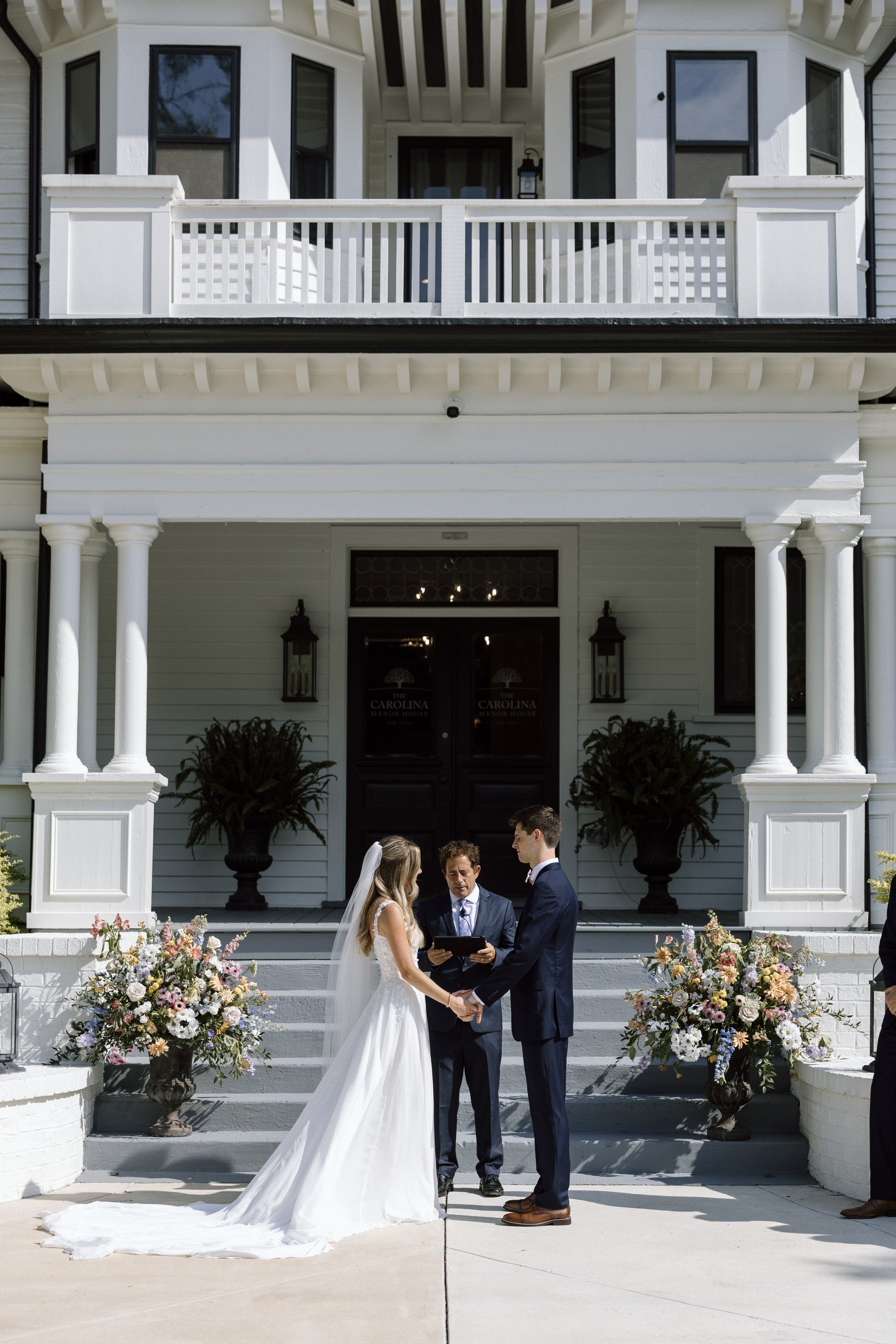 Couple getting married outside a white house with floral arrangements and an officiant.
