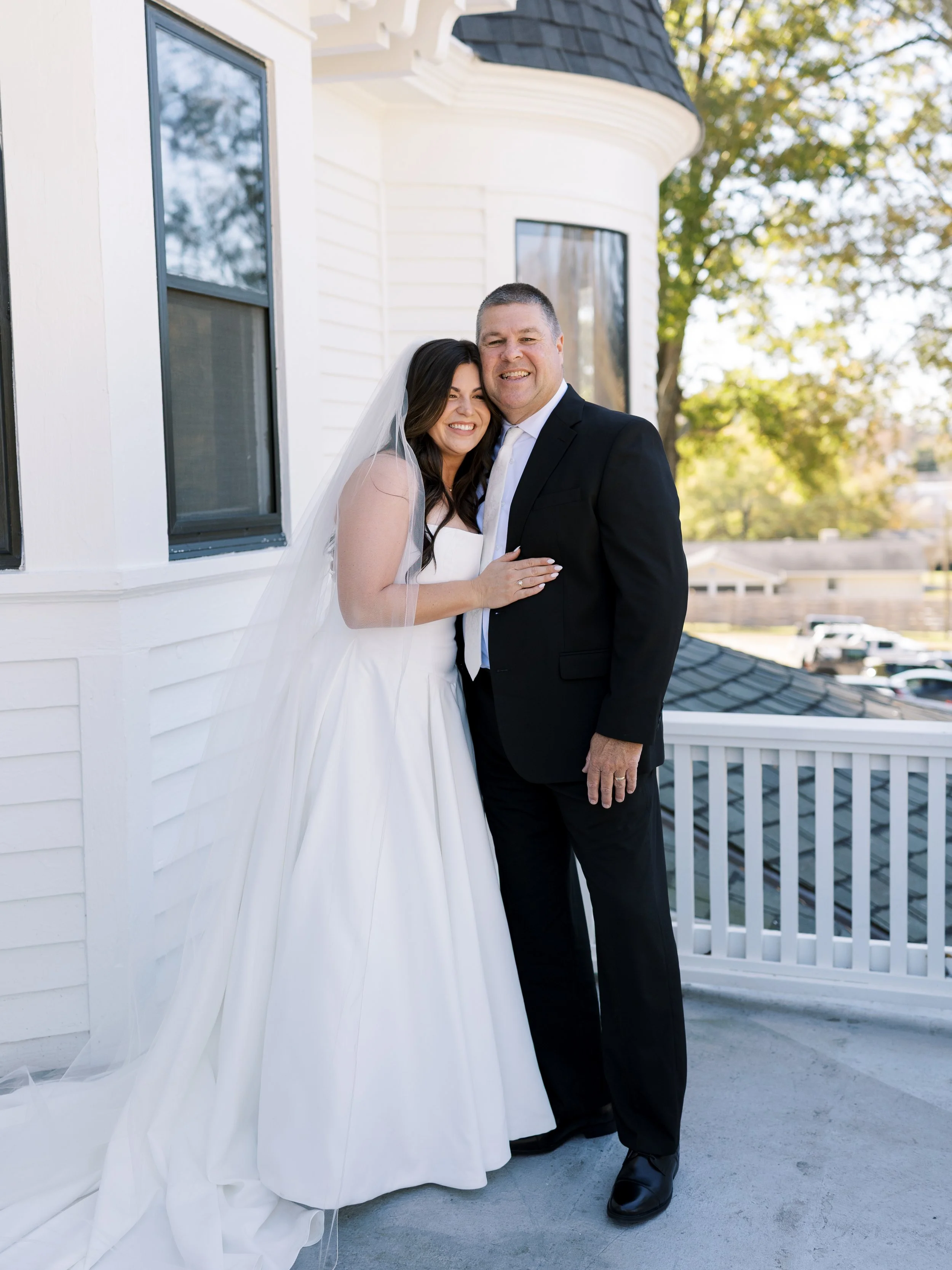 Bride and groom standing close together on a balcony, smiling, with the bride in a white wedding gown and veil, and the groom in a black suit, outside a white house with trees in the background.