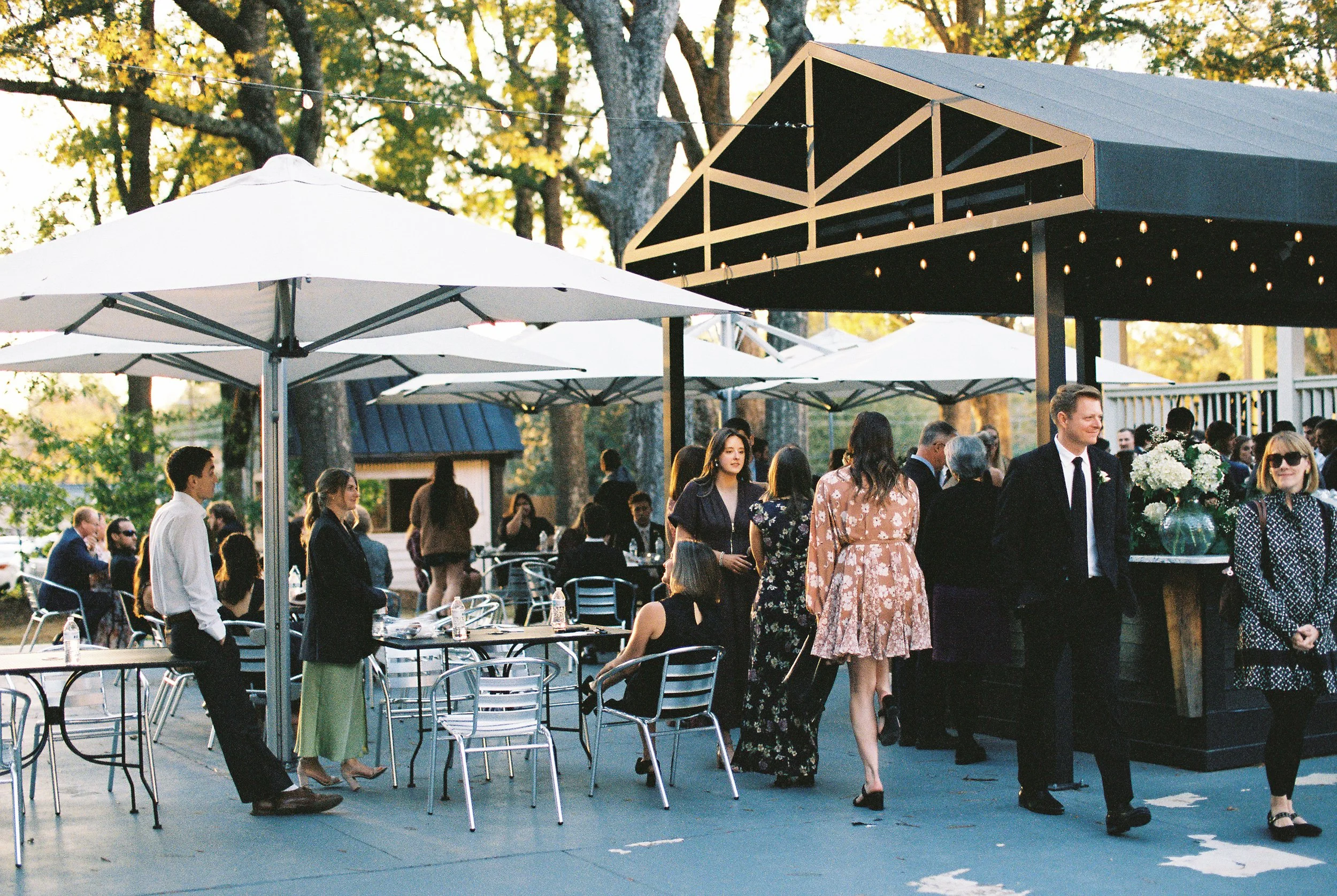 People gathered at an outdoor event with tables, chairs, and umbrellas, under a shelter decorated with string lights and surrounded by trees.