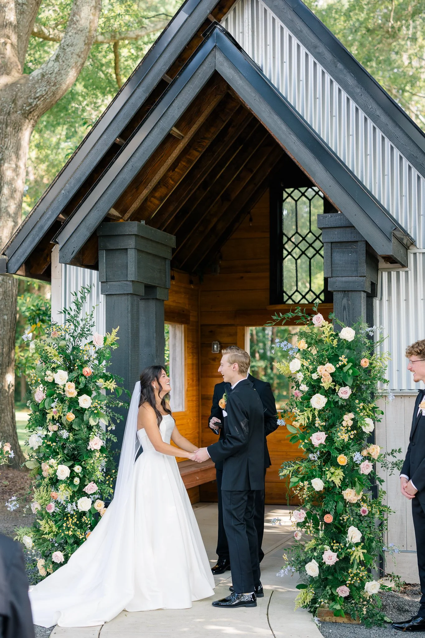 Bride and groom exchanging vows under a floral arch at an outdoor wedding ceremony.