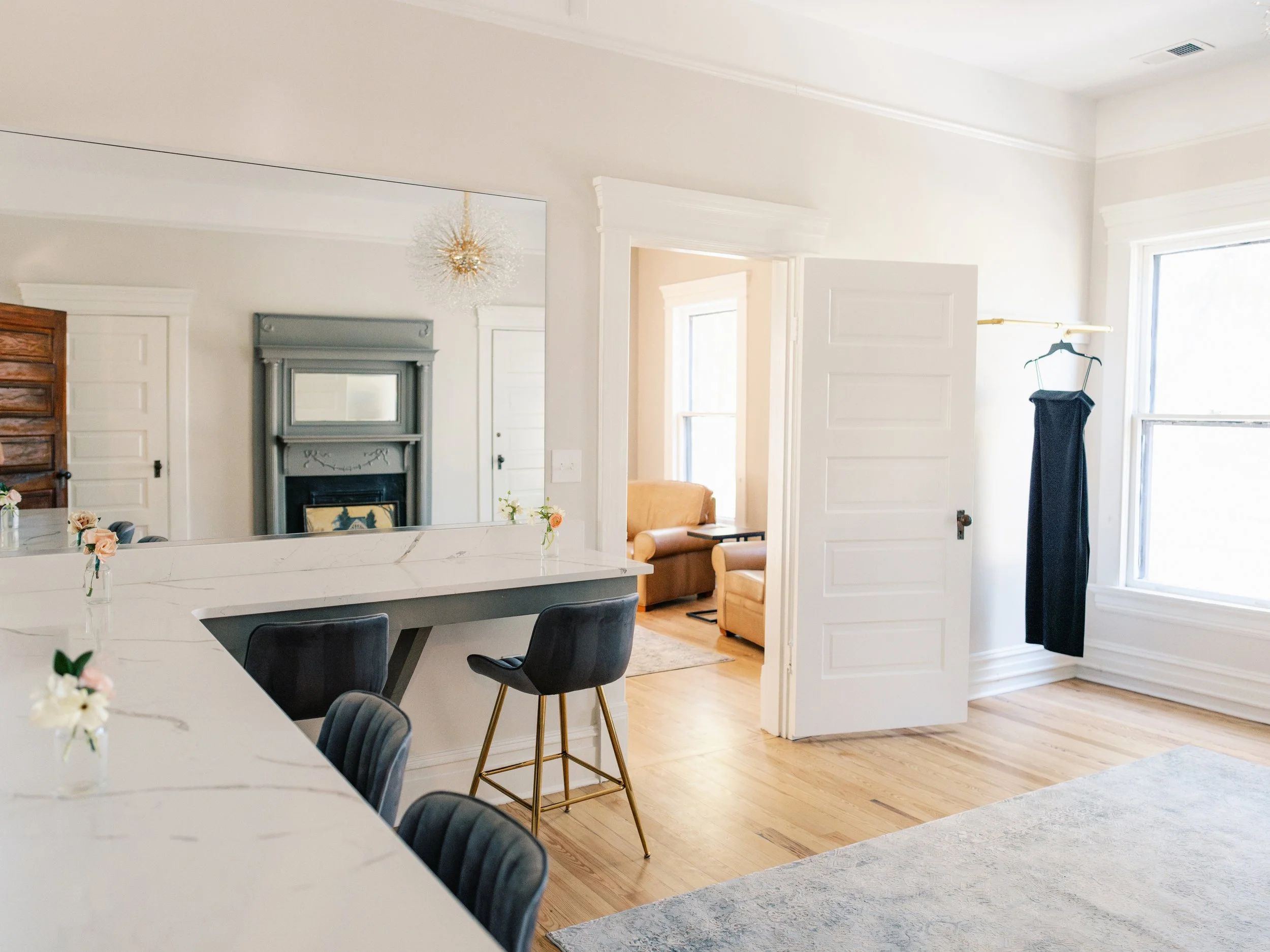 A bright dining and living area with large windows, hardwood floors, a white marble kitchen island with black chairs, a mirror without a frame, a corner with a wardrobe and a hanging black dress, and a view into a sitting room with a fireplace and tan sofa.