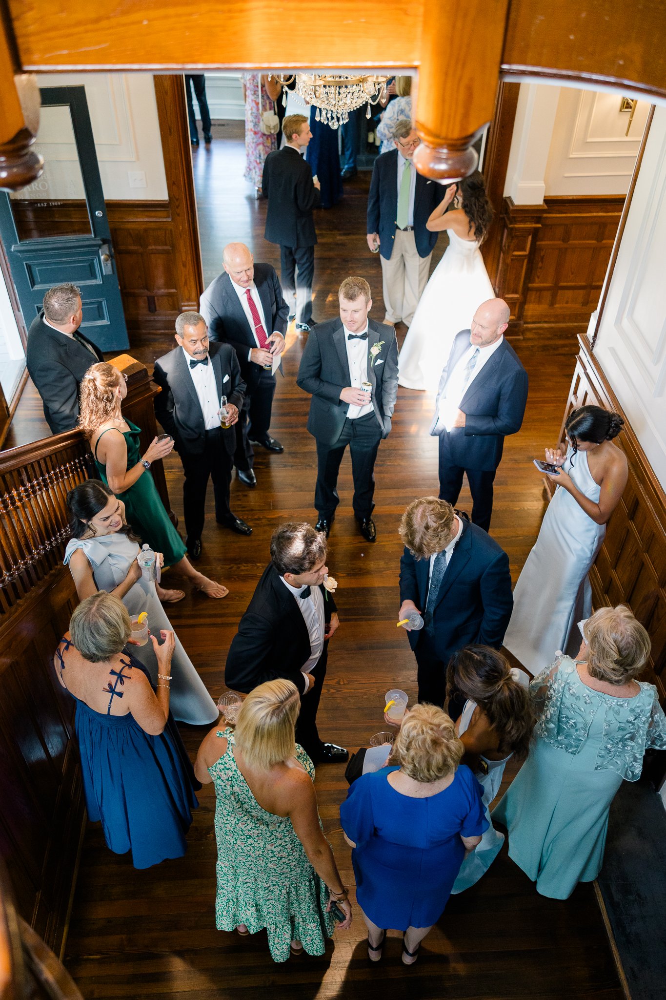 A wedding reception with guests socializing on a wooden-floored upper level, some holding drinks, with a bride and groom partly visible in the background.