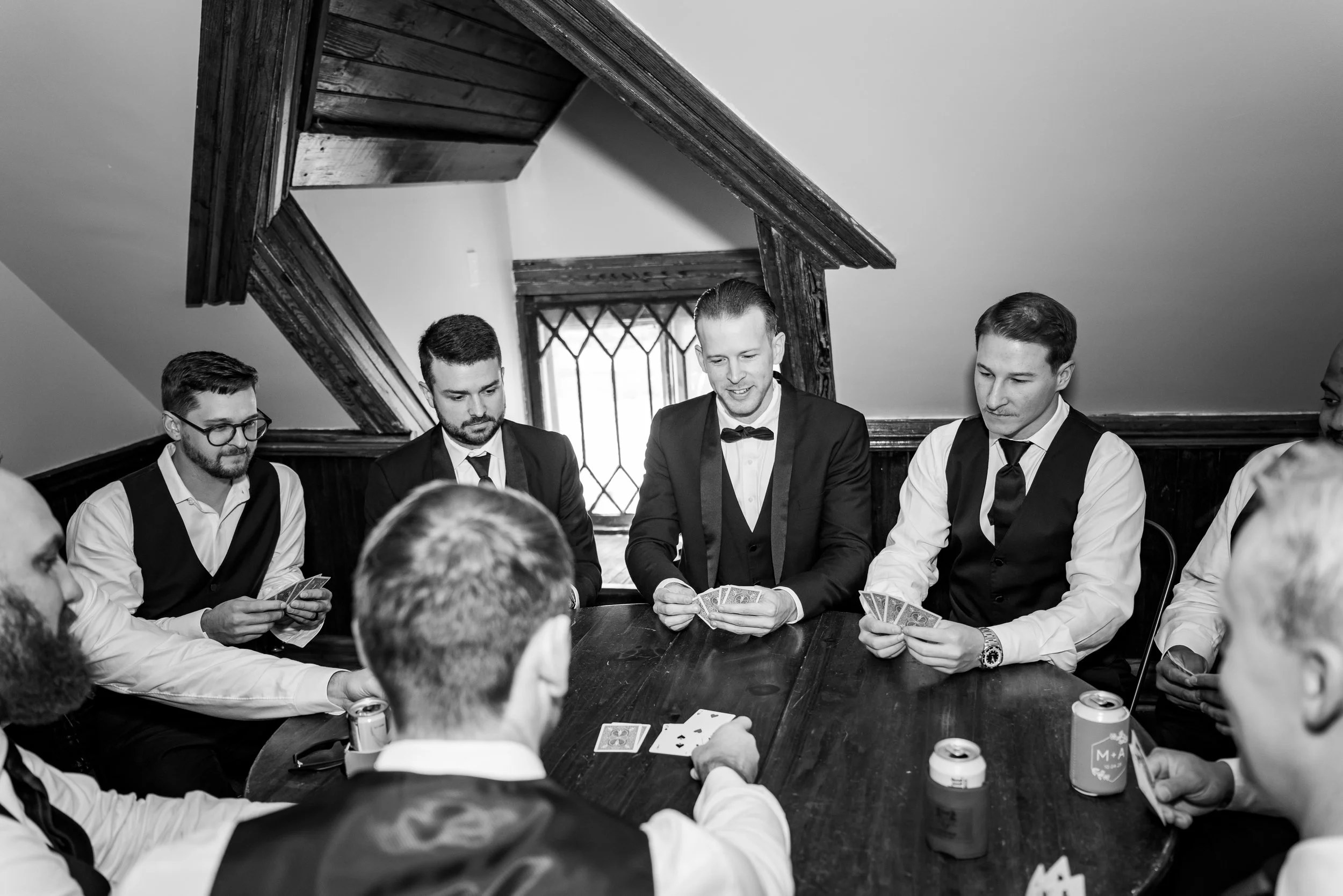 Black-and-white photo of a group of men in suits and vests playing cards around a wooden table in a room with wood-paneled walls and a small diamond window.