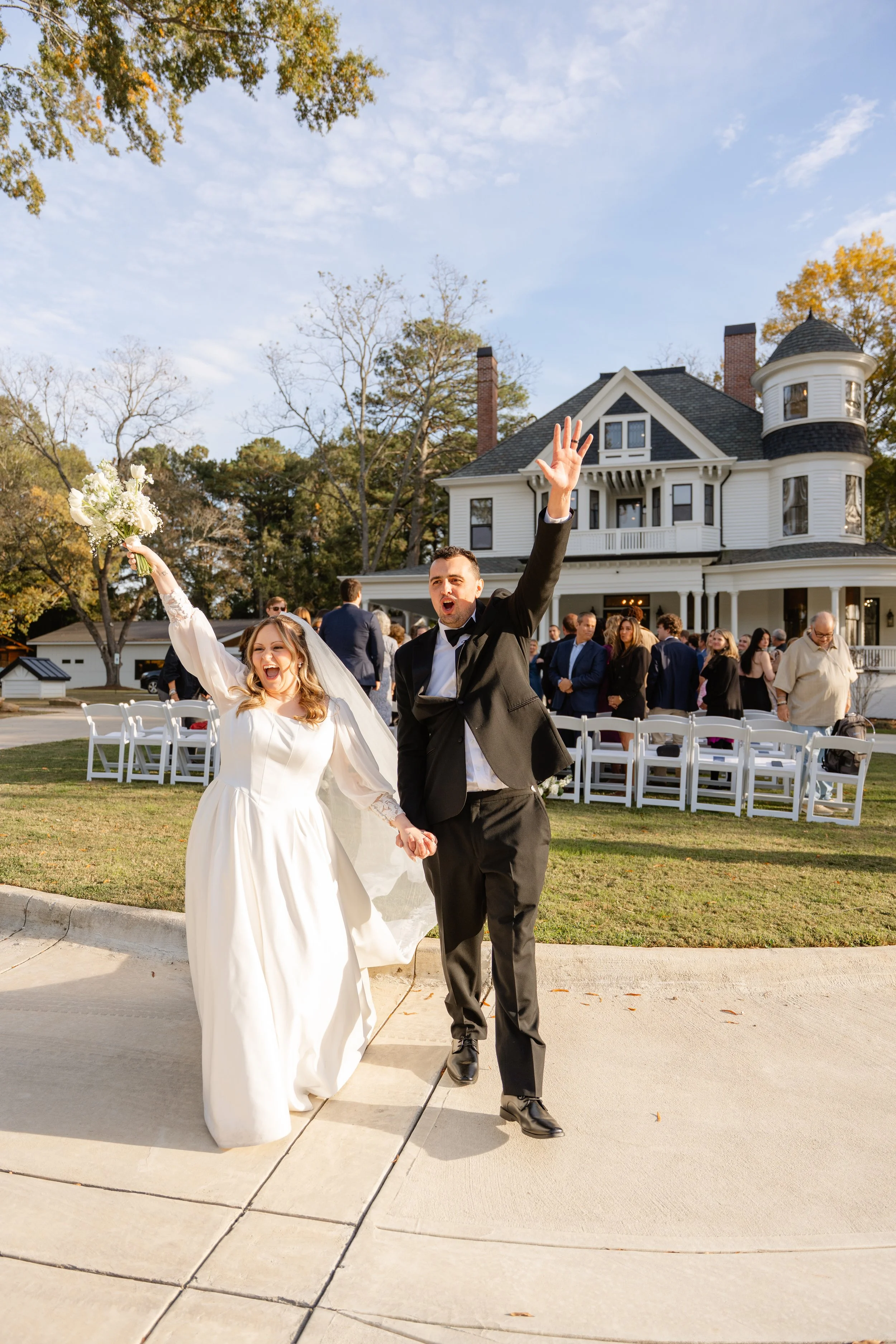 A newlywed couple celebrating their wedding outside a large, historic white house. The bride is wearing a long white dress and holding a bouquet, while the groom is in a black tuxedo. They are joyful, walking hand in hand with the bride raising her b