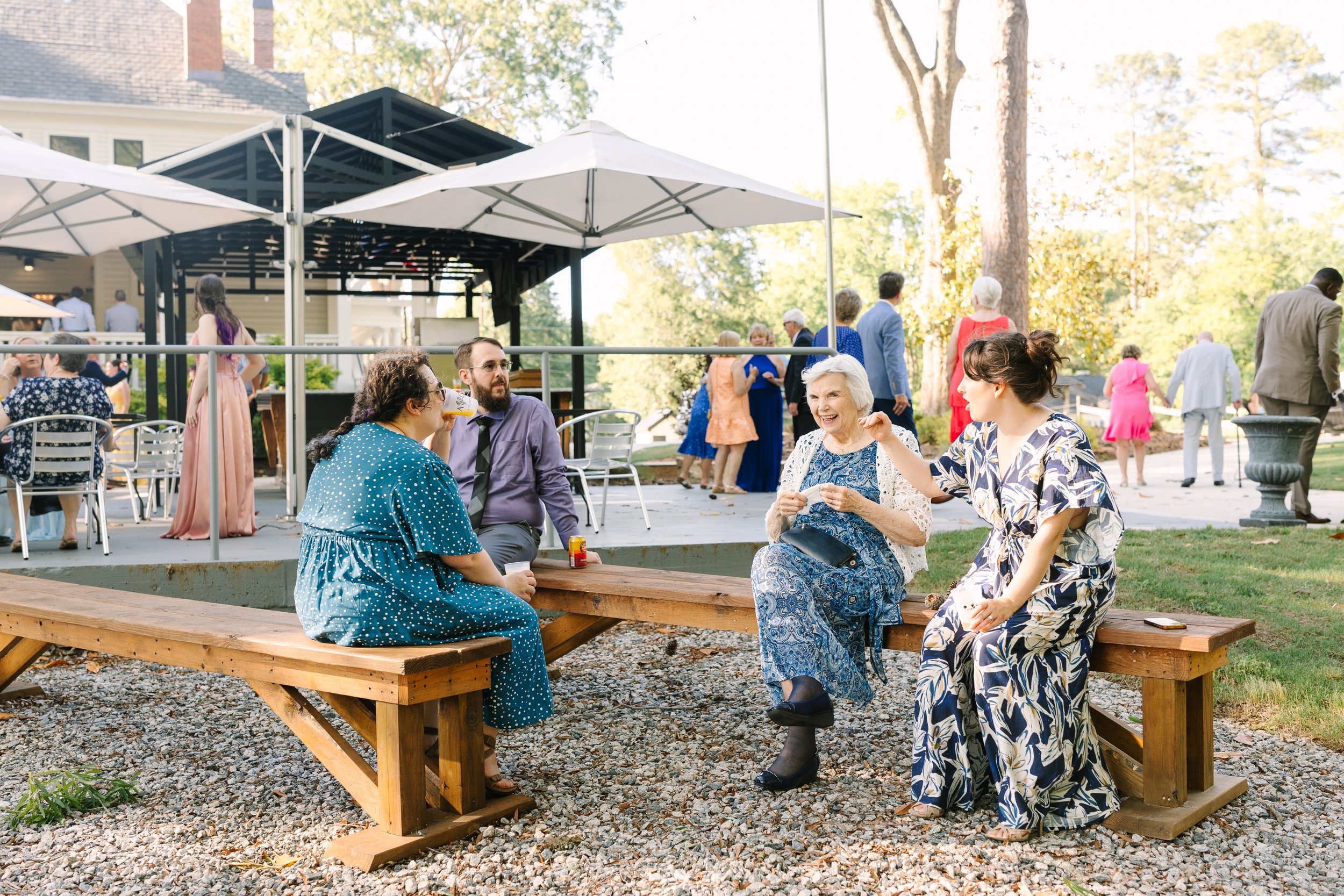 People socializing at an outdoor gathering, sitting on wooden benches and standing under a canopy, with trees and a building in the background.