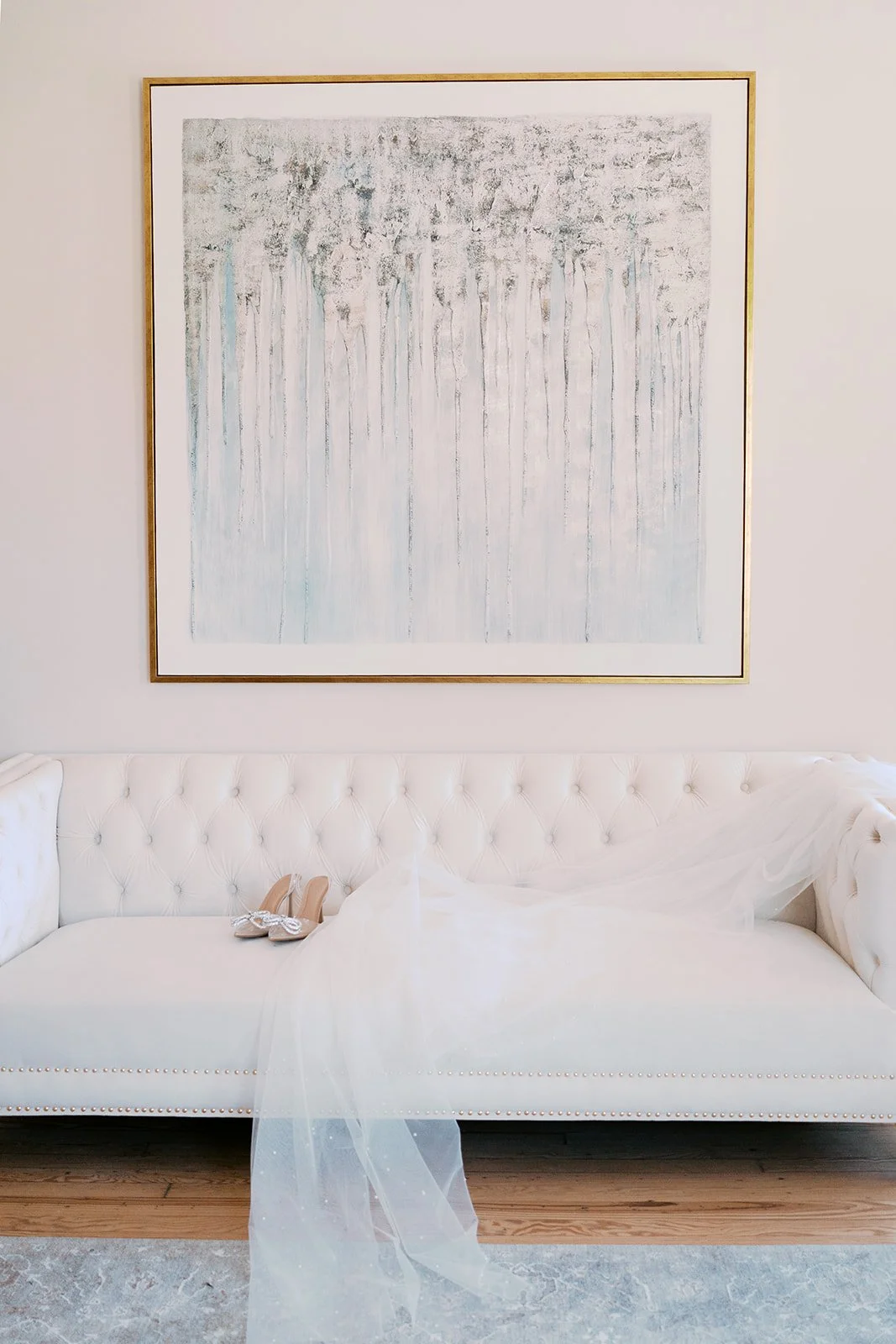 A white tufted sofa with a pair of beige high-heeled shoes and a white veil or fabric draped over it, positioned below a large abstract painting of trees on a white wall.