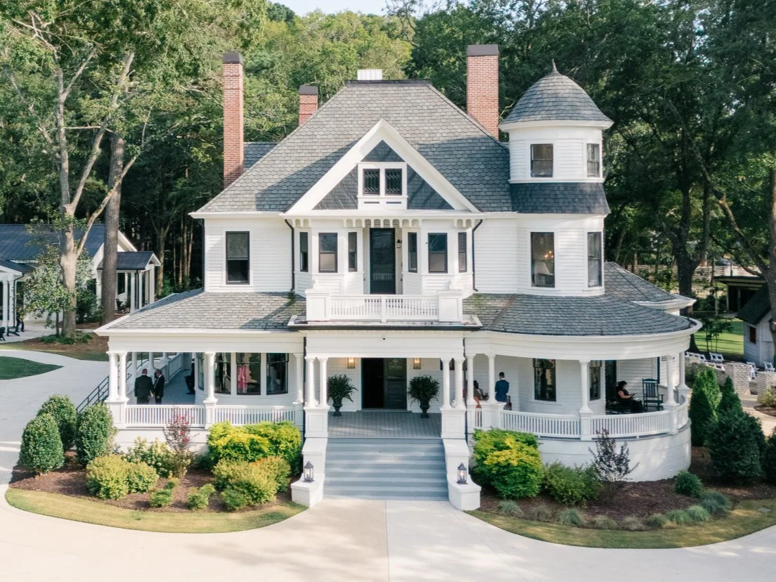 Large white Victorian-style house with a wraparound porch, turret on the right, multiple chimneys, surrounded by trees and landscaped yard.