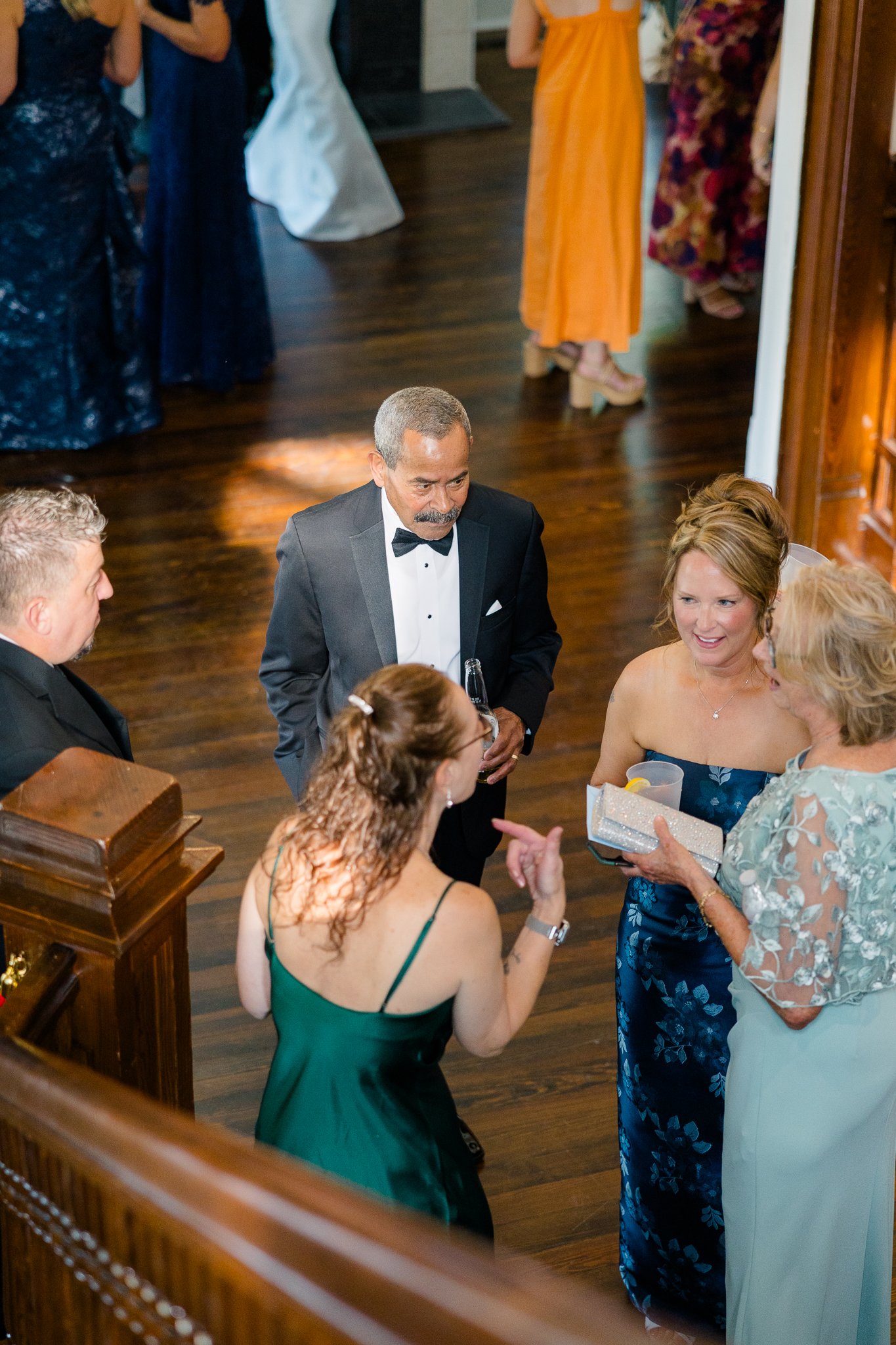 Group of people at a formal event, including a man in a tuxedo and women in elegant dresses, engaged in conversation on a wooden floor.