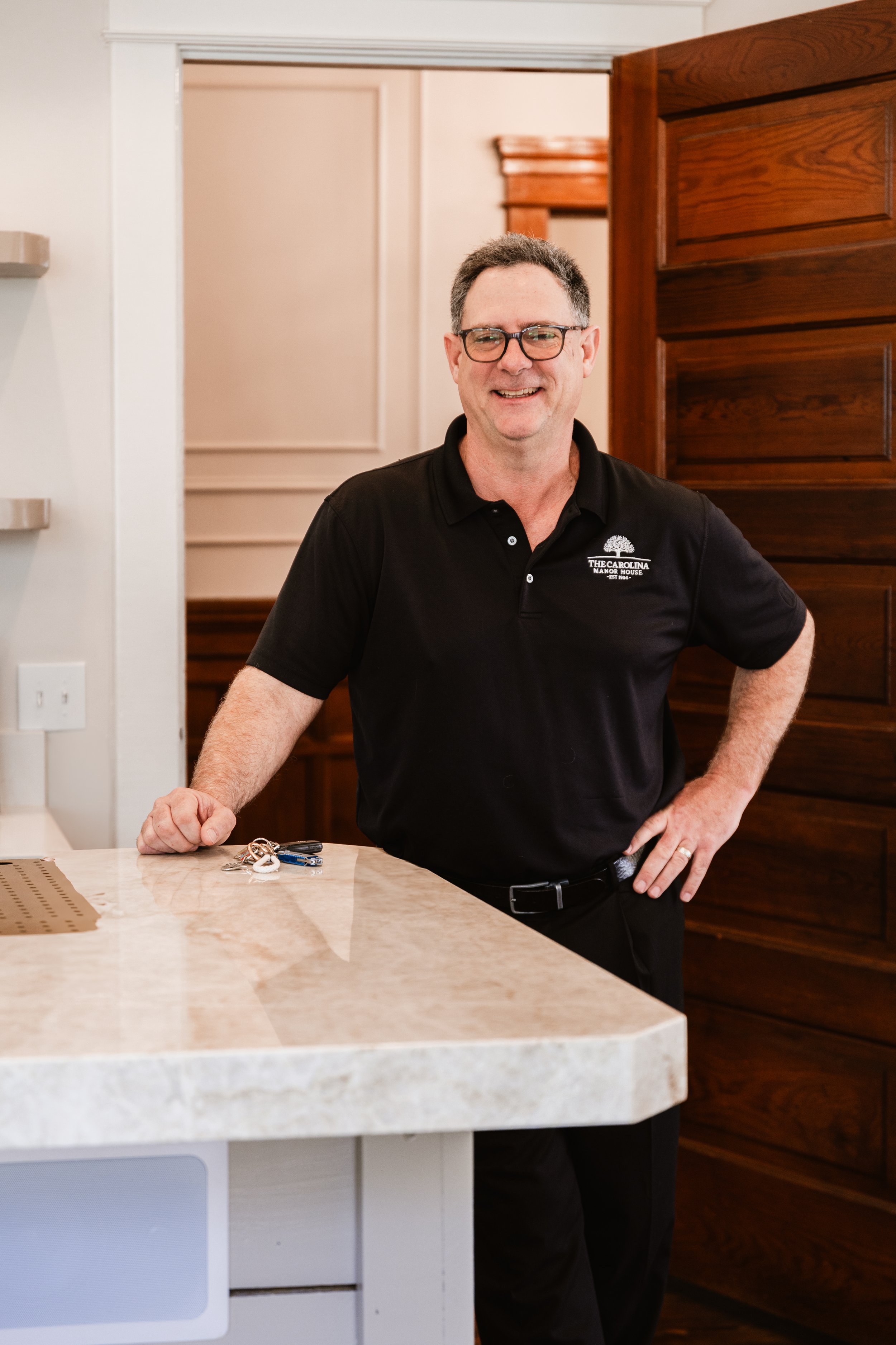 A man wearing glasses and a black polo shirt with a logo stands at a kitchen counter, smiling and posing for the camera.