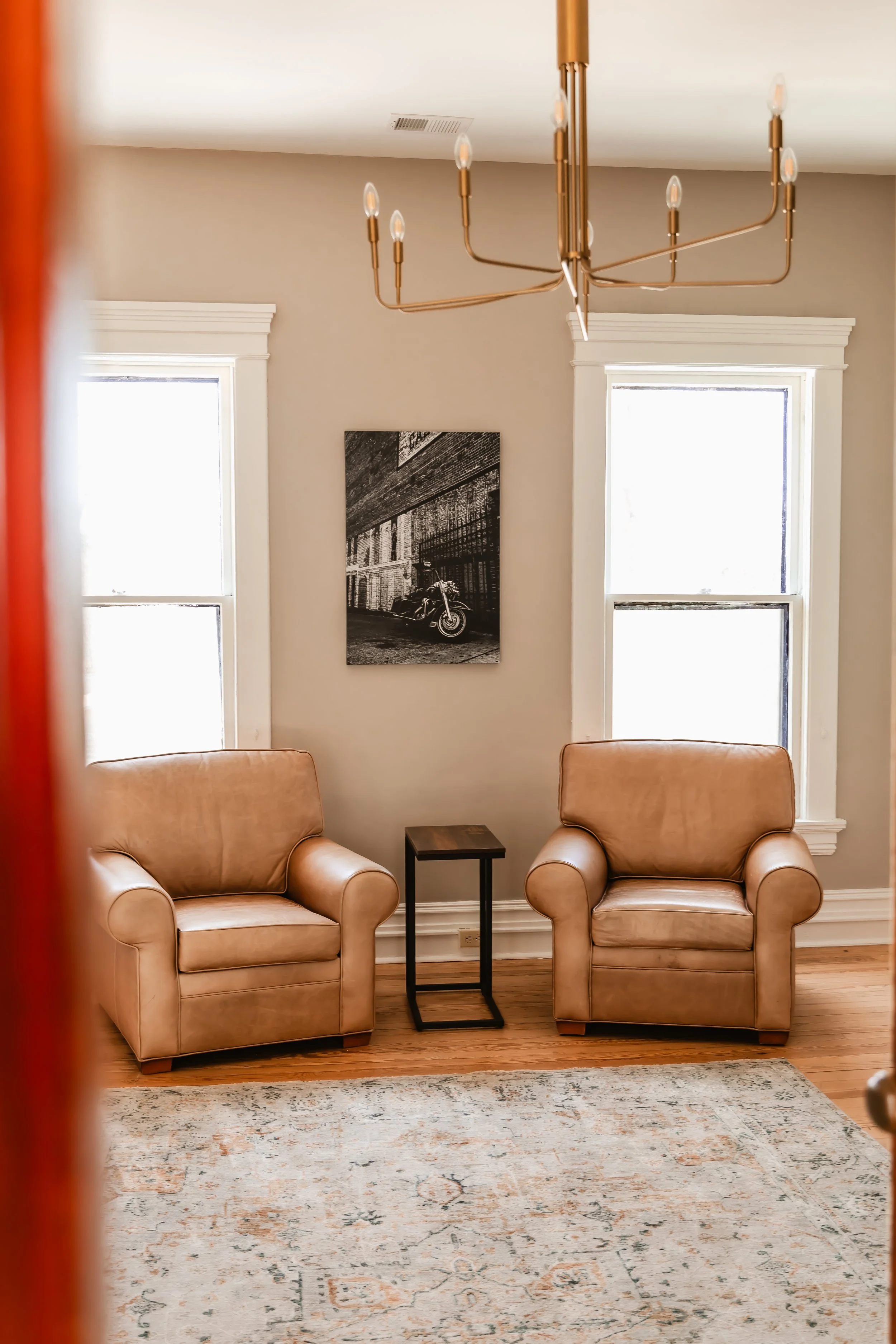 Living room with two beige armchairs, a small black side table between them, a black and white photo on the wall, two large windows, and a gold chandelier hanging from the ceiling.