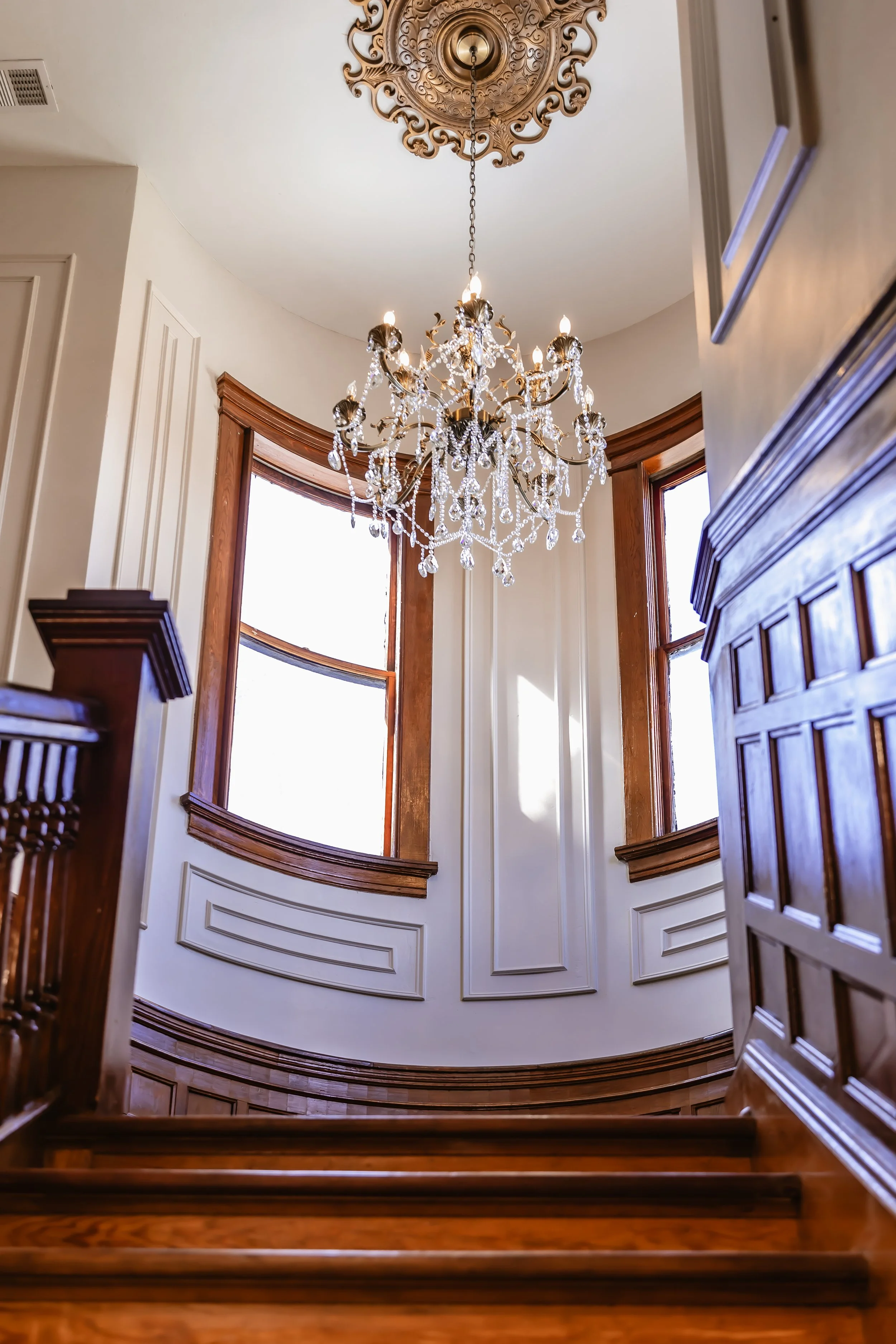 Interior view of a staircase area with large windows, a chandelier hanging from the ceiling, and wood trim around the windows and staircase.