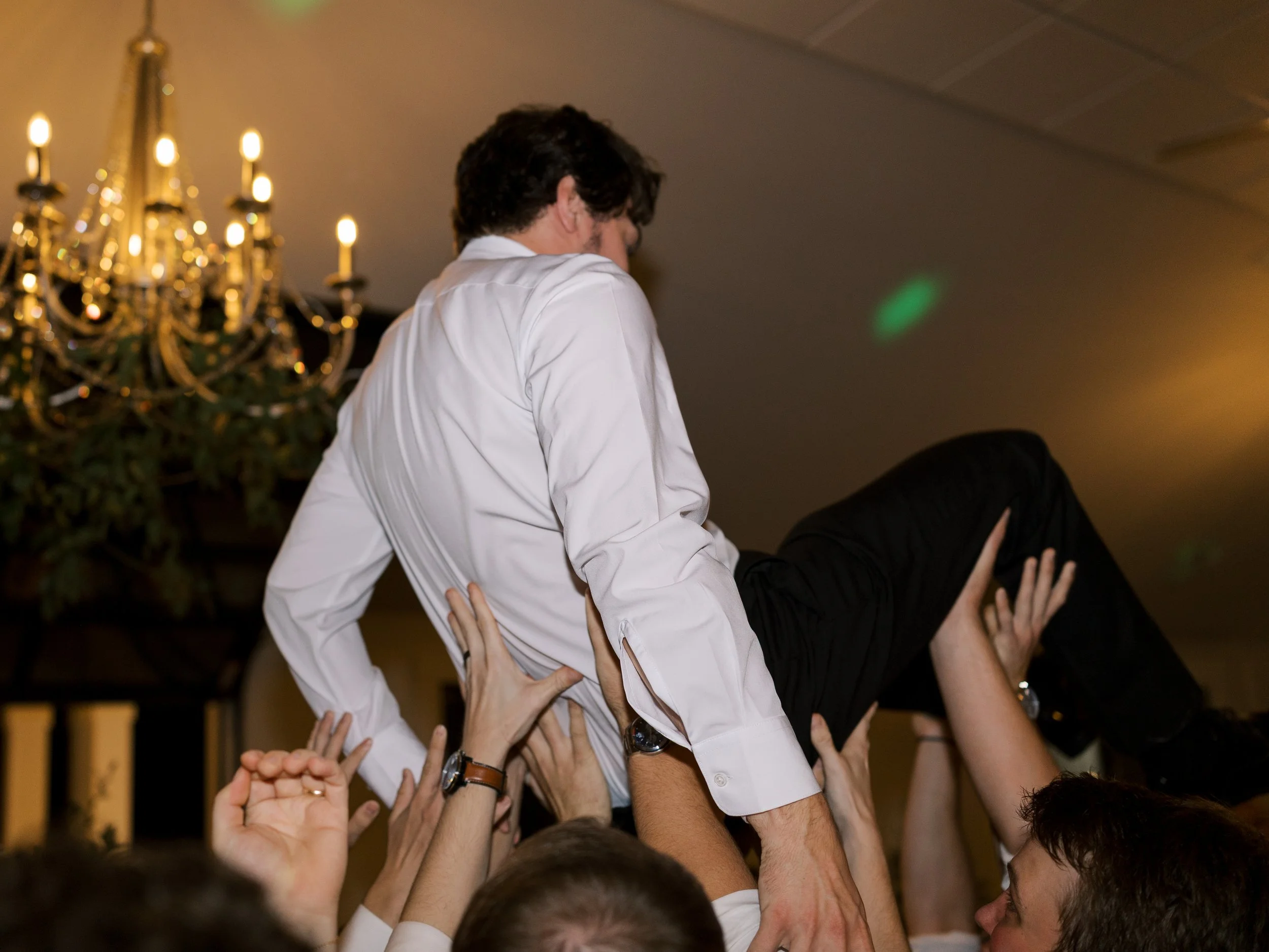 A man being lifted on a crowd's hands at a celebration event, with a chandelier visible in the background.