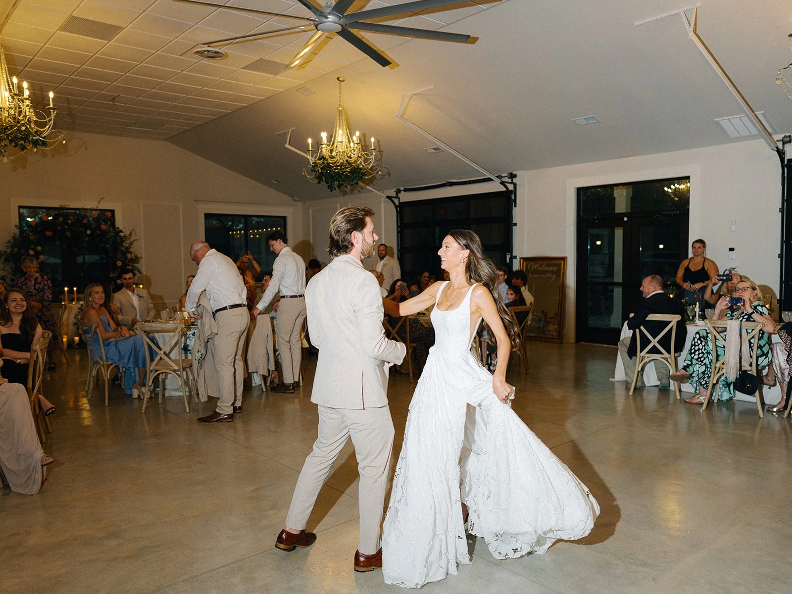 A bride and groom are dancing at their wedding reception, surrounded by seated guests and waitstaff in a decorated hall with chandeliers, ceiling fans, and large windows.