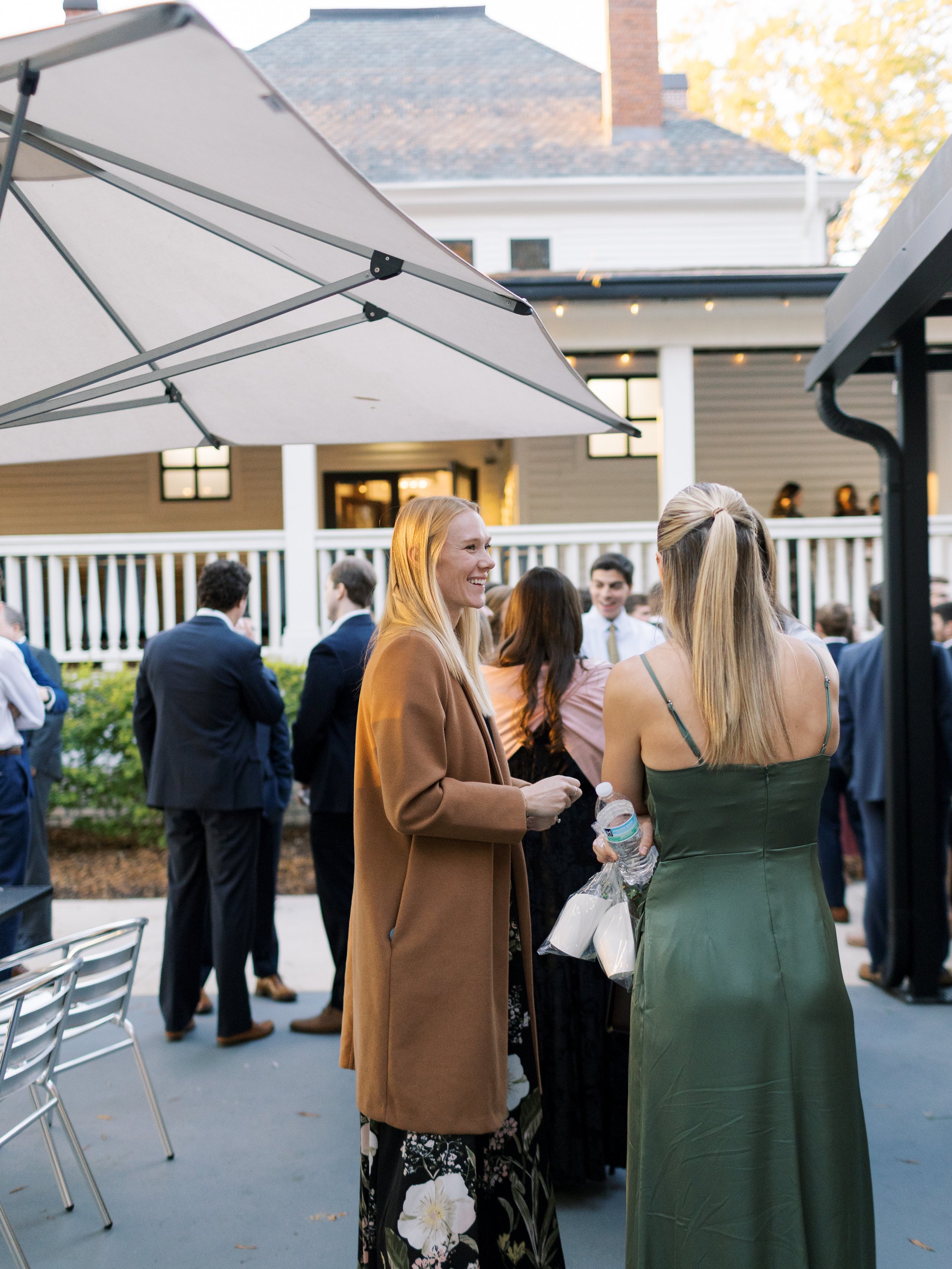 A social outdoor gathering with people chatting, dressed in semi-formal attire, on a patio with an umbrella, in front of a house with a porch and string lights.