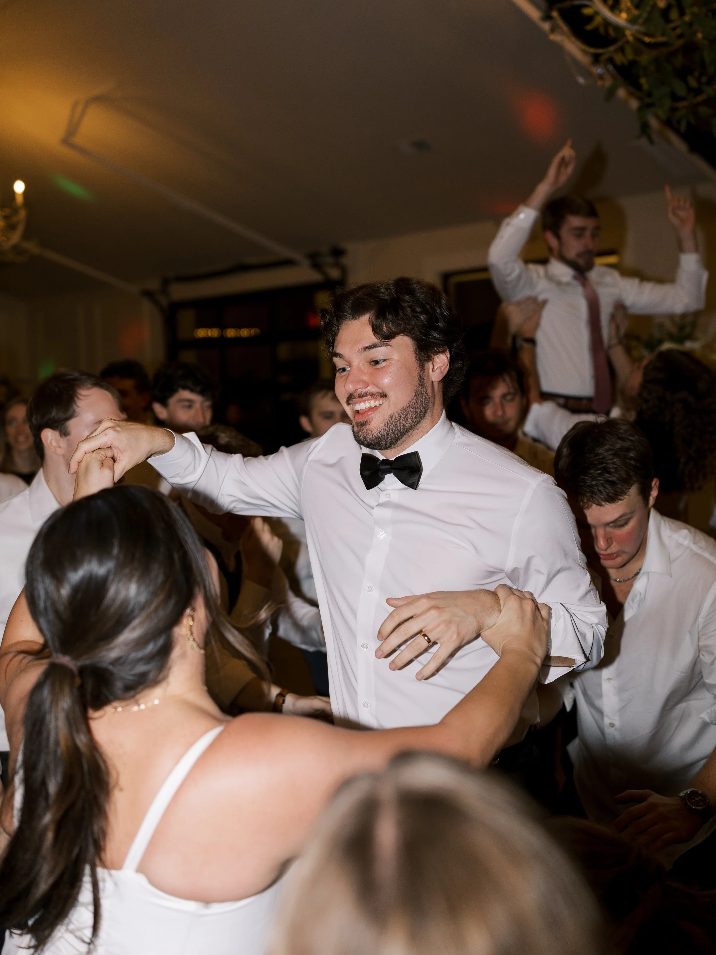 Man in tuxedo dancing at a lively indoor celebration with guests surrounding him, some dancing and some watching, with warm lighting and a festive atmosphere.