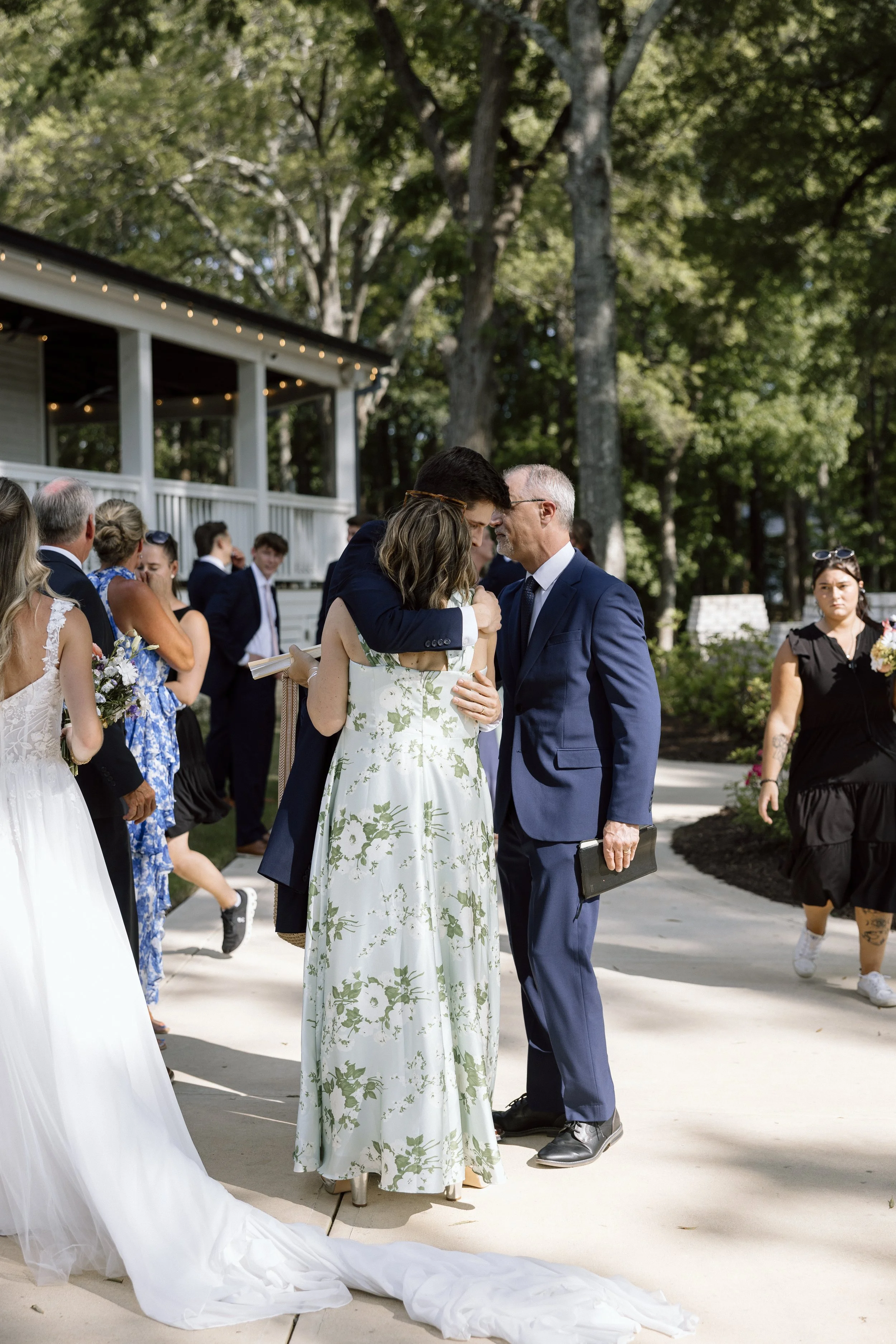 A group of people at an outdoor wedding, with two individuals sharing an emotional moment in the center as they embrace, surrounded by guests and trees.