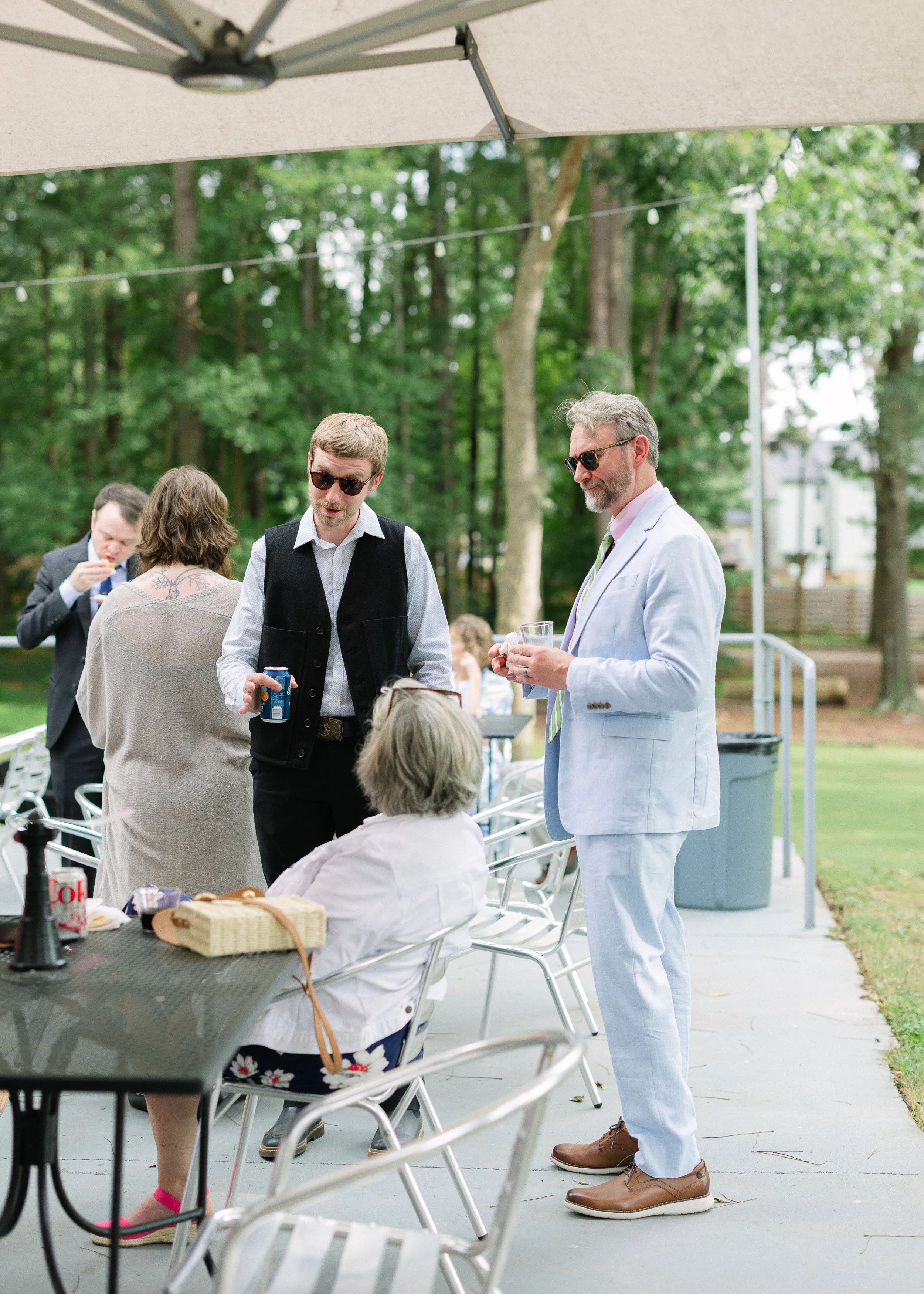People socializing outdoors at a gathering under a canopy, with trees in the background, seated around a table with drinks and snacks.
