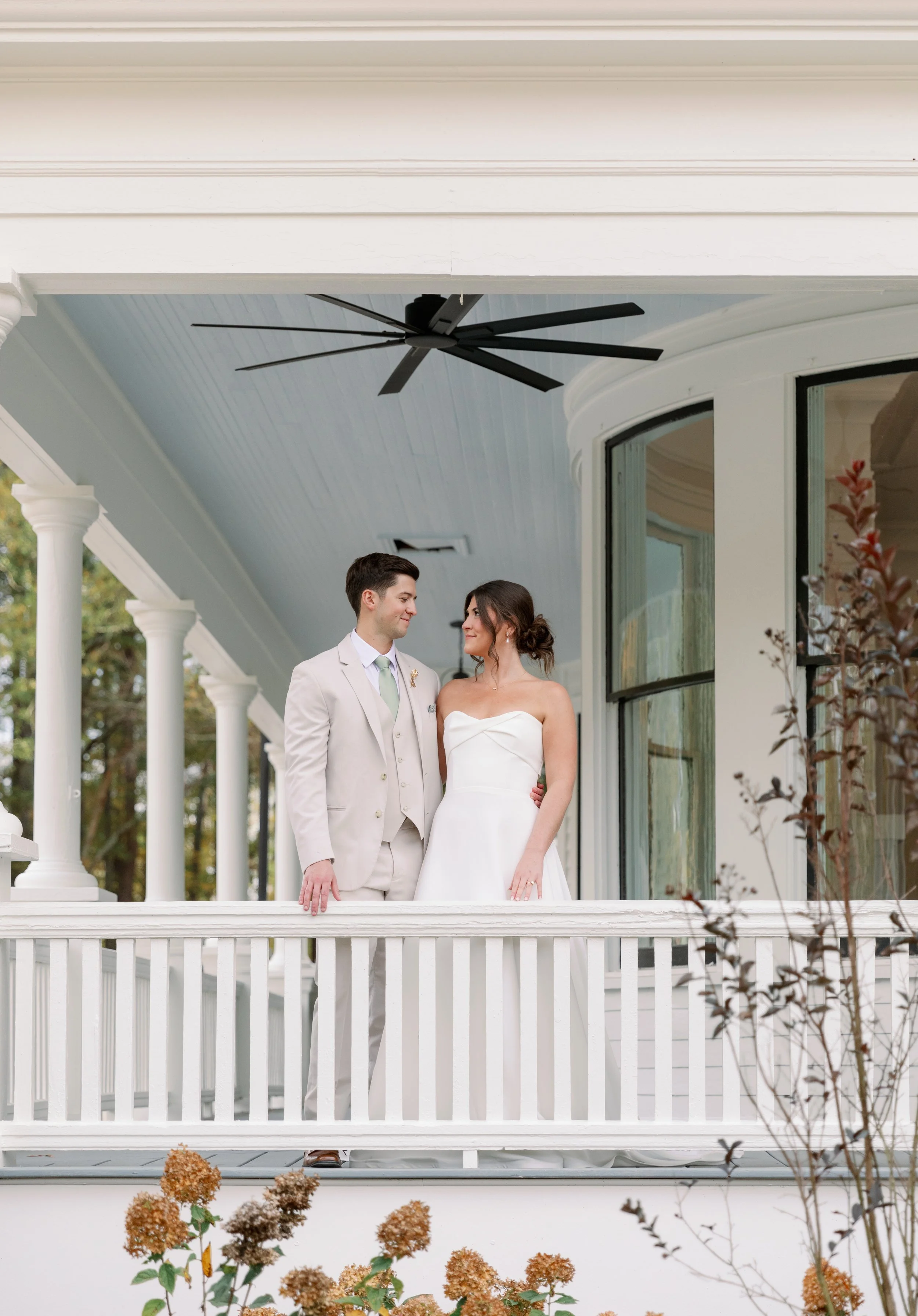 A bride and groom standing on a porch, looking at each other, with a white railing and a house behind them, decorated with plants and trees.