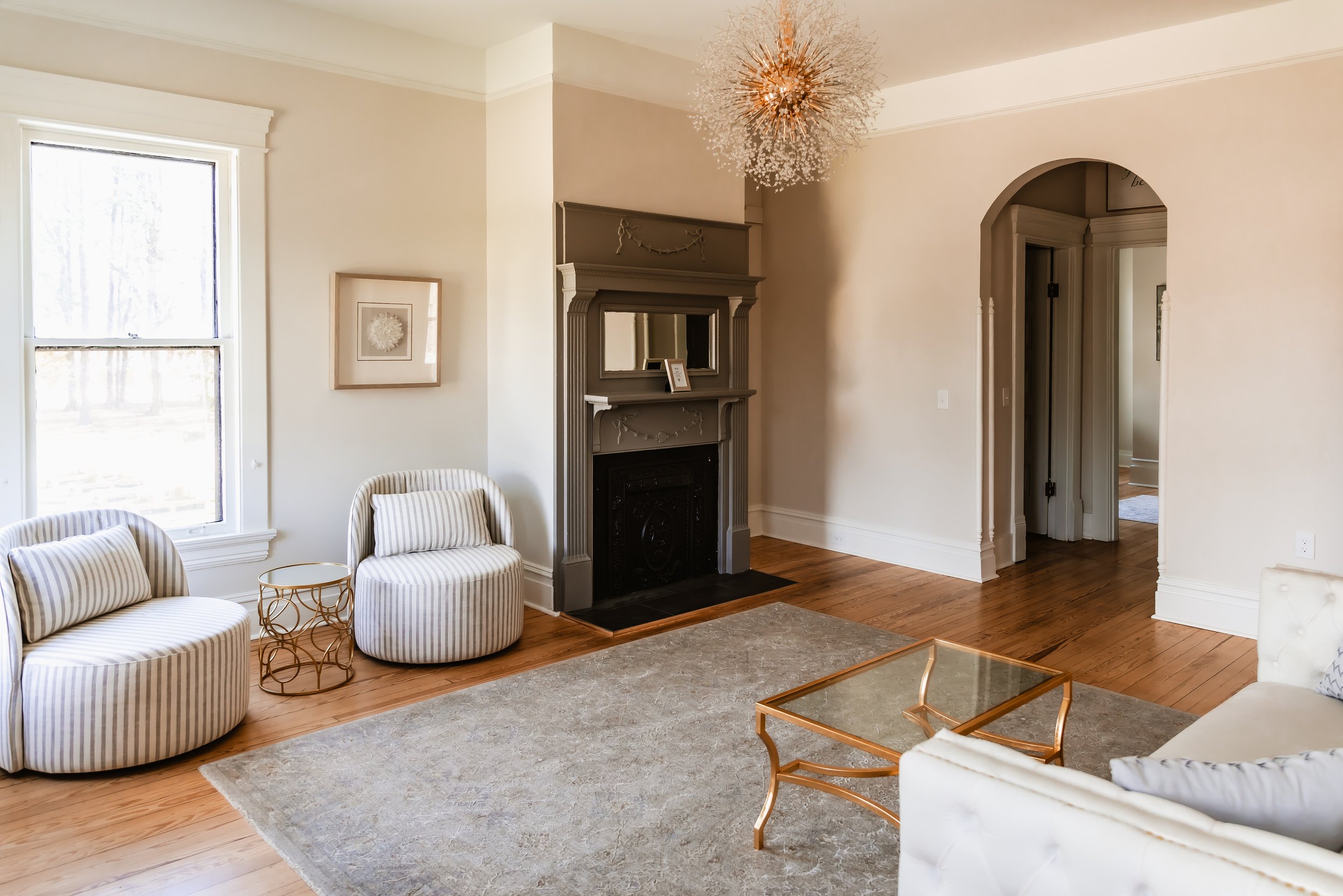 Living room with two striped armchairs, a beige sofa, a gold coffee table, a fireplace, a large window, a framed picture, and a modern chandelier.