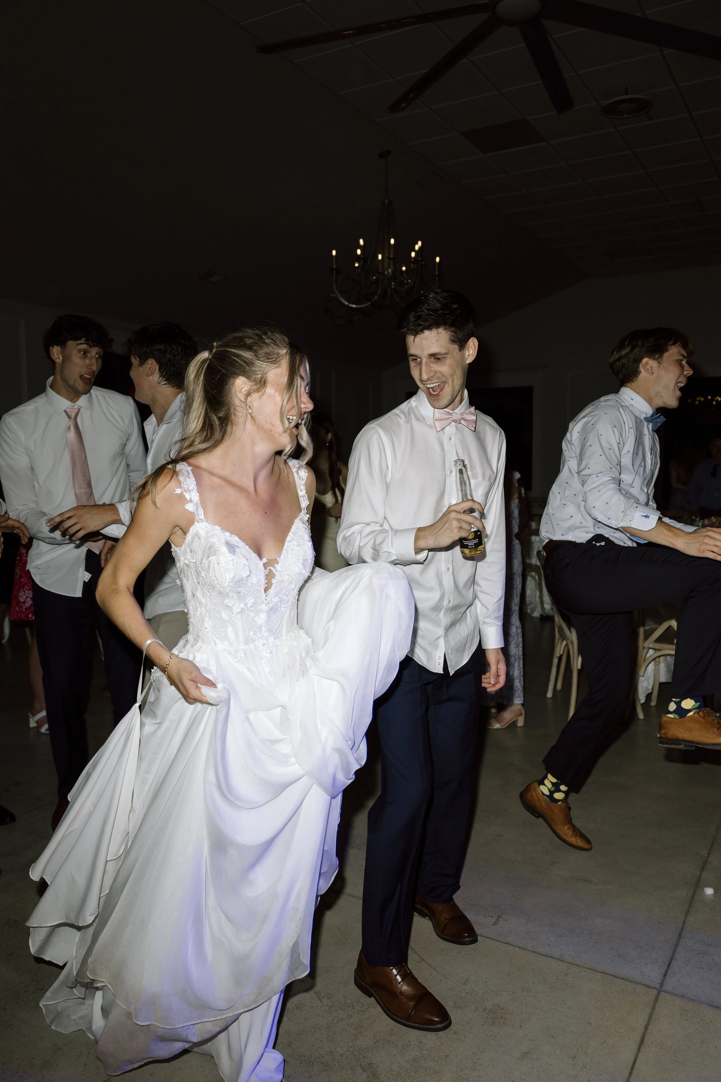At a wedding reception, a bride lifting her dress revealing her leg, walking with a man holding a bottle, while others dance and socialize in the background.