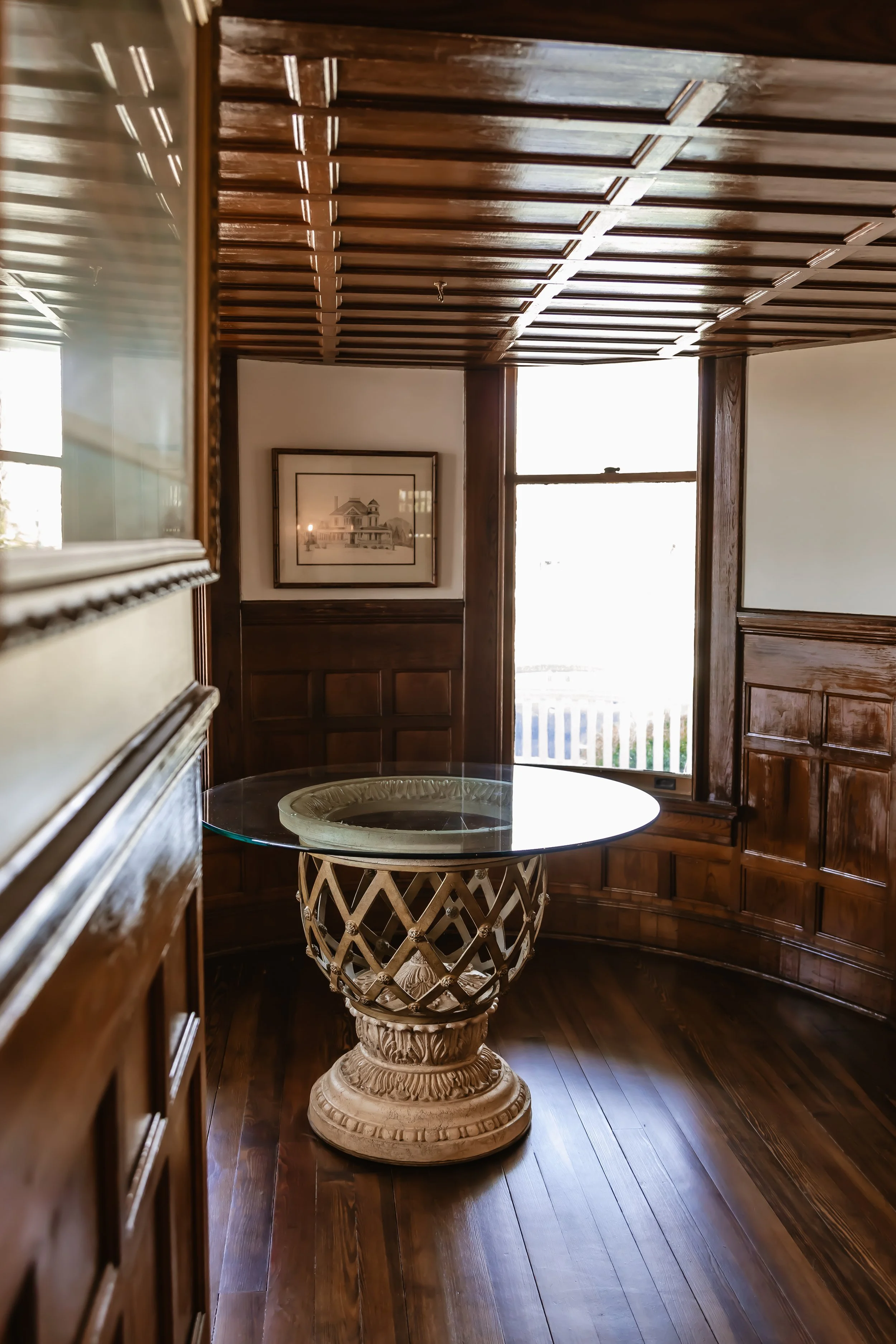 A corner of a room with dark wooden paneled walls and ceiling, a large window with sunlight, a framed picture of a house on the wall, and a round table with a glass top and ornate carved base.