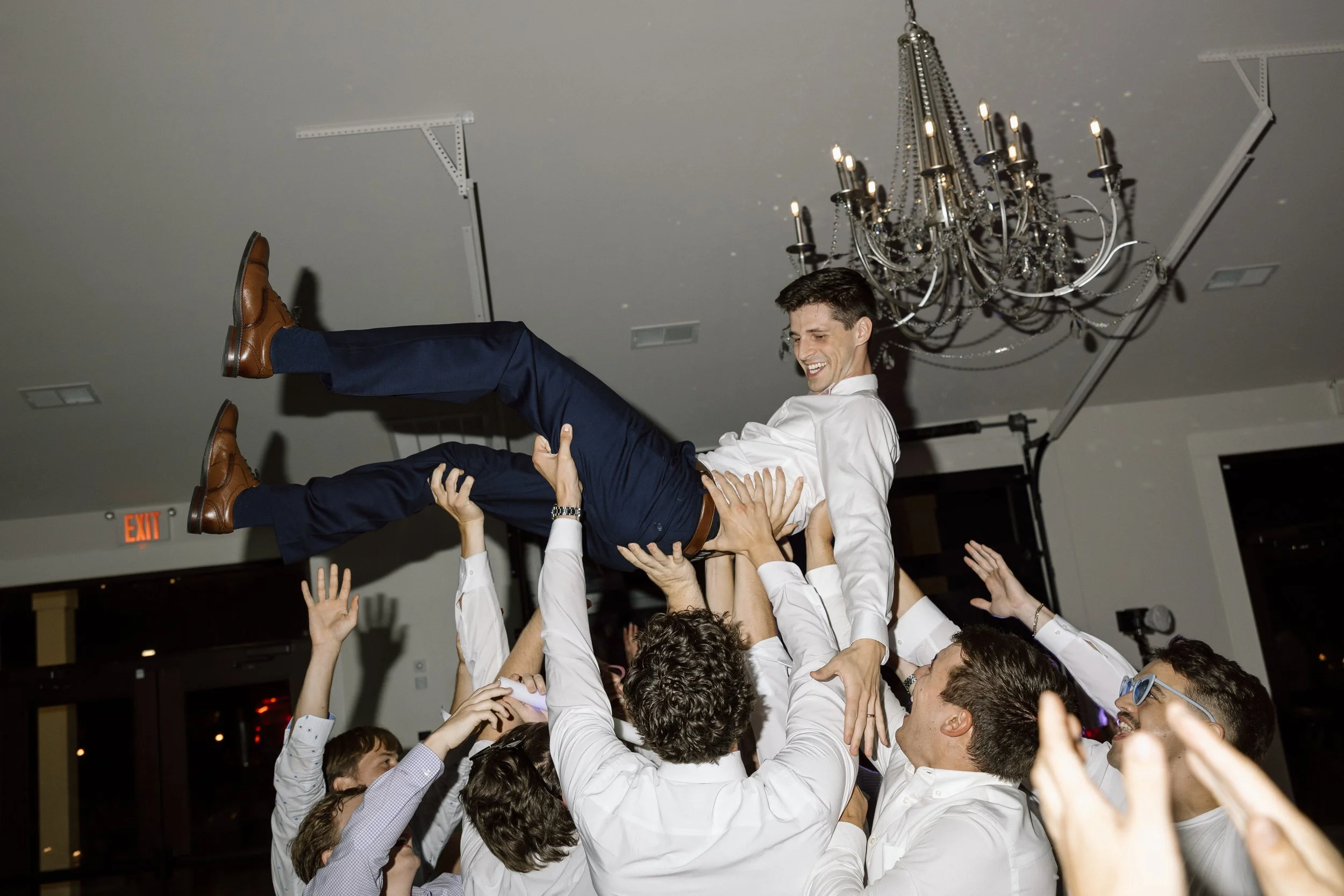 A group of people celebrating at a party, lifting a man in a white shirt and dark blue pants into the air. The man is smiling and wearing brown shoes, his arms are outstretched, and everyone appears joyful. The scene is indoors, with a chandelier han