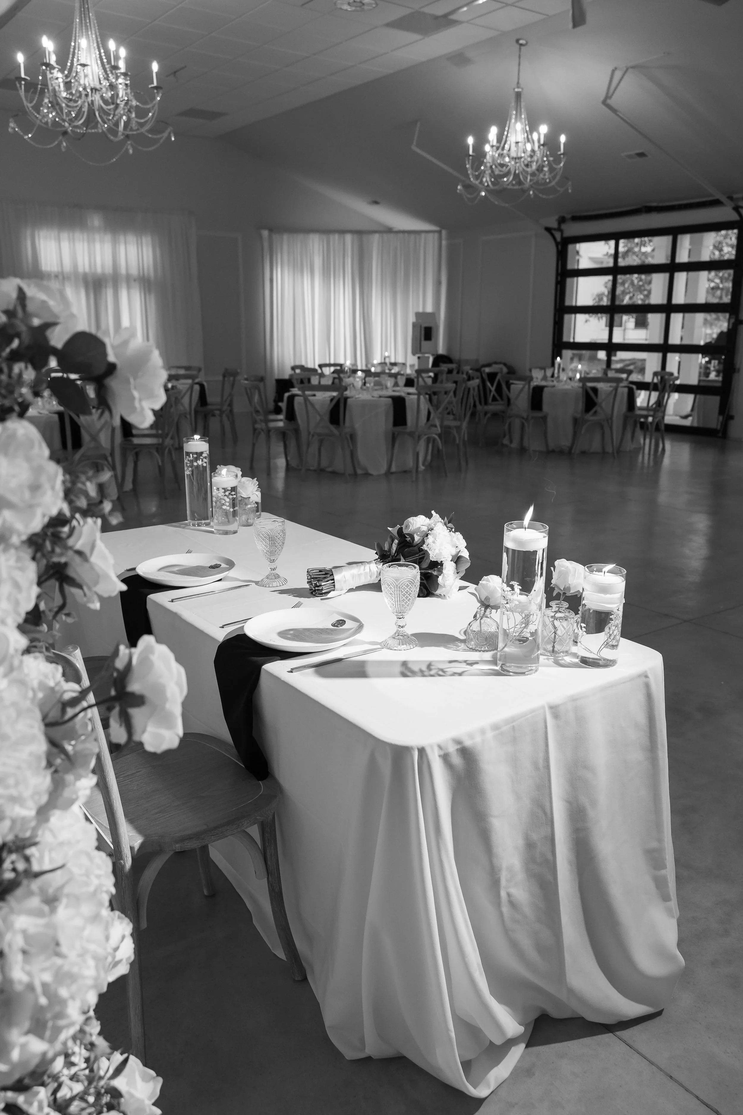 Elegant event venue with decorated tables, floral arrangements, candles, and chandeliers in black and white photograph.
