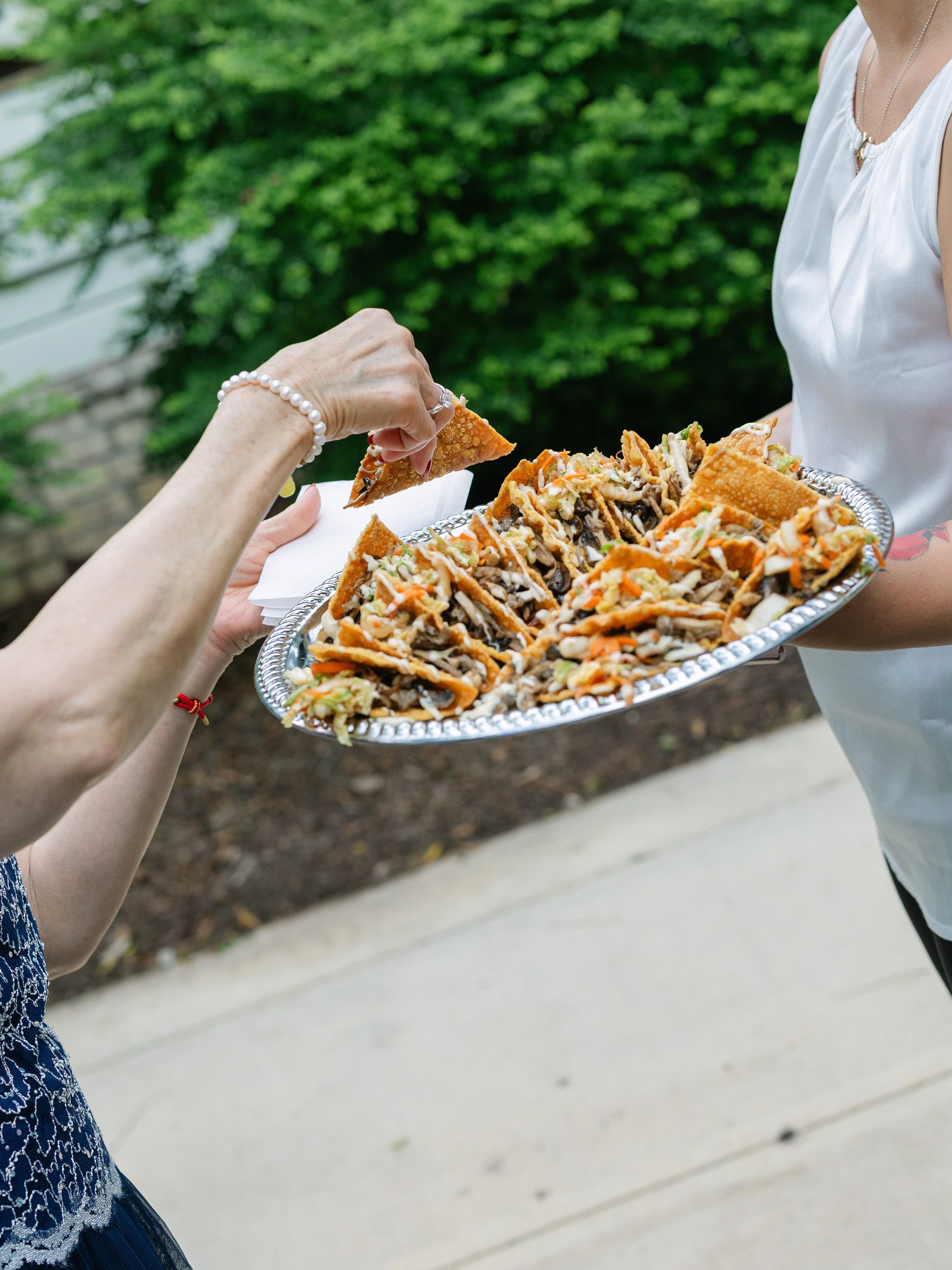 Person serving tacos to another person outdoors with green foliage in the background.