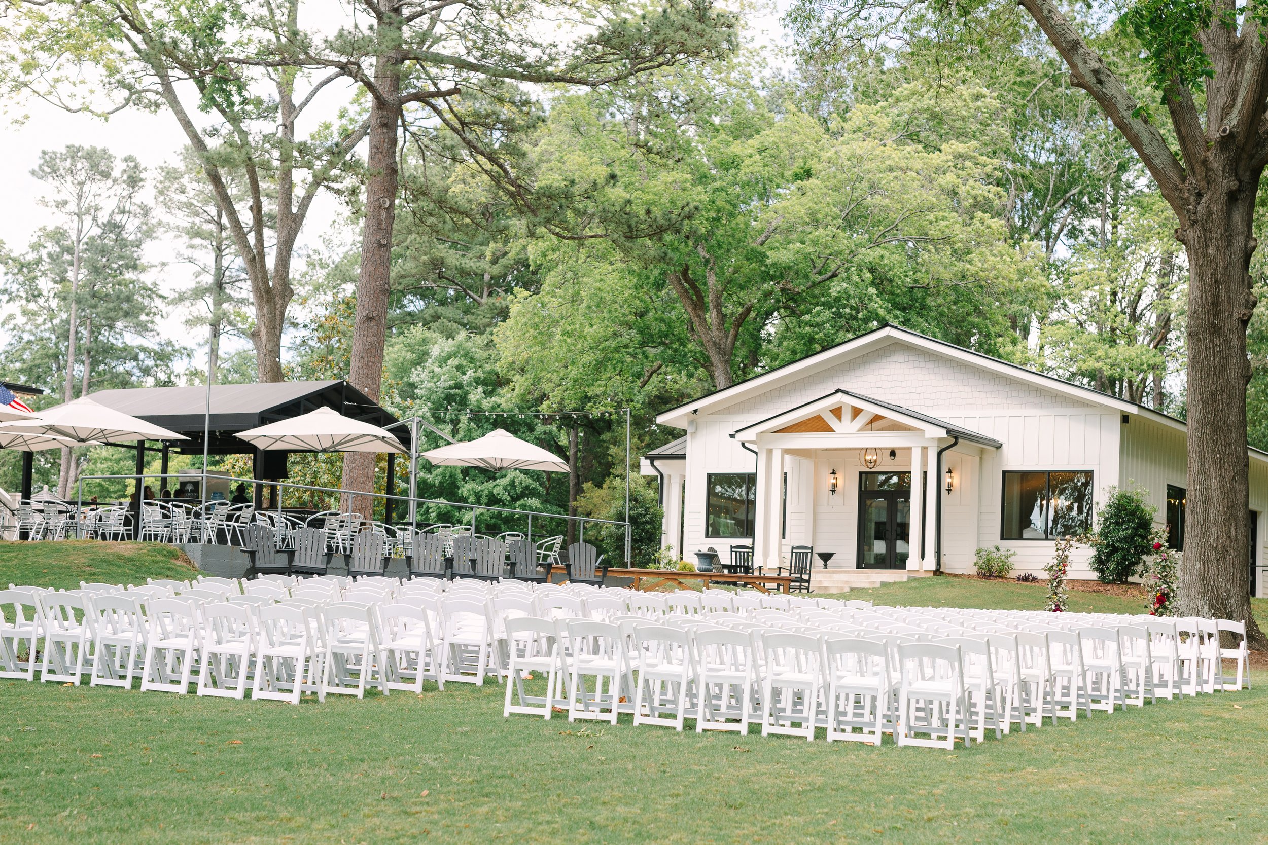 Outdoor wedding setup with dozens of white chairs arranged in front of a white chapel, surrounded by trees and outdoor umbrellas at a scenic venue.