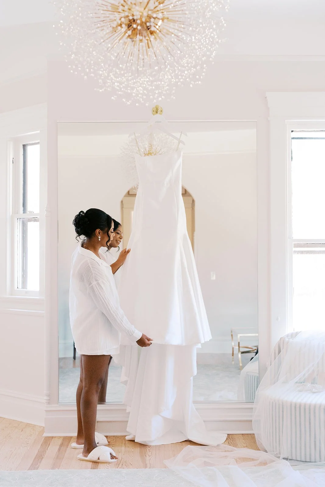 Two women looking at a hanging white wedding dress in a brightly lit room with a large mirror and windows.