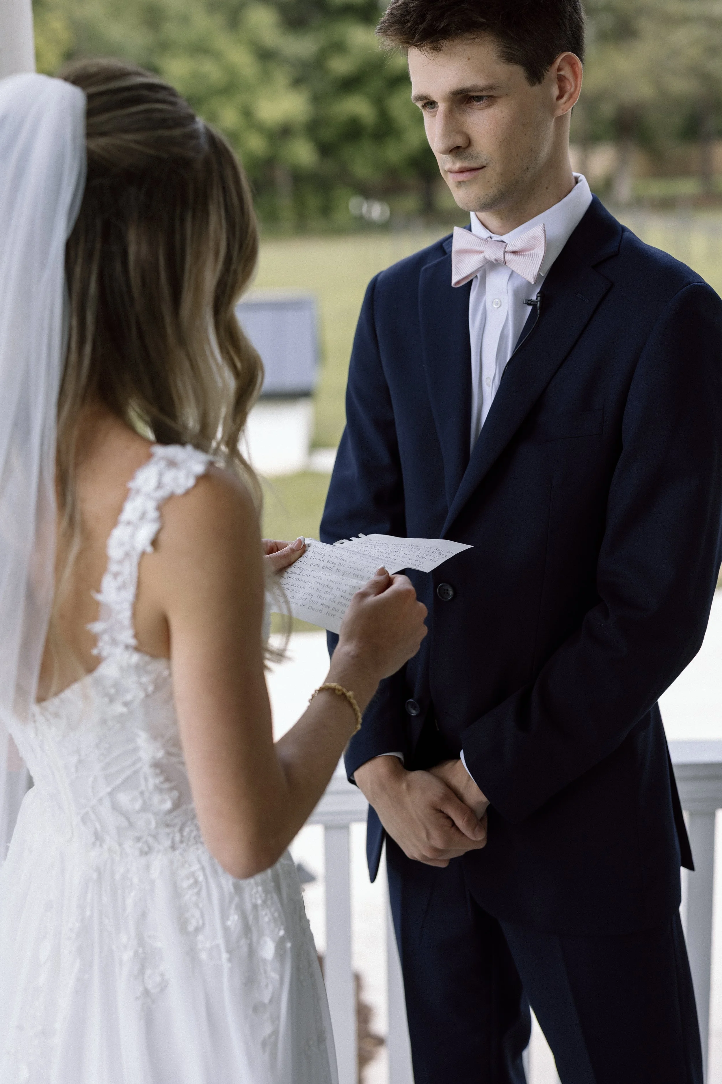 A bride and groom during wedding vows, with the bride in a white gown with lace details and the groom in a dark suit with a pink bow tie, exchanging vows outdoors.