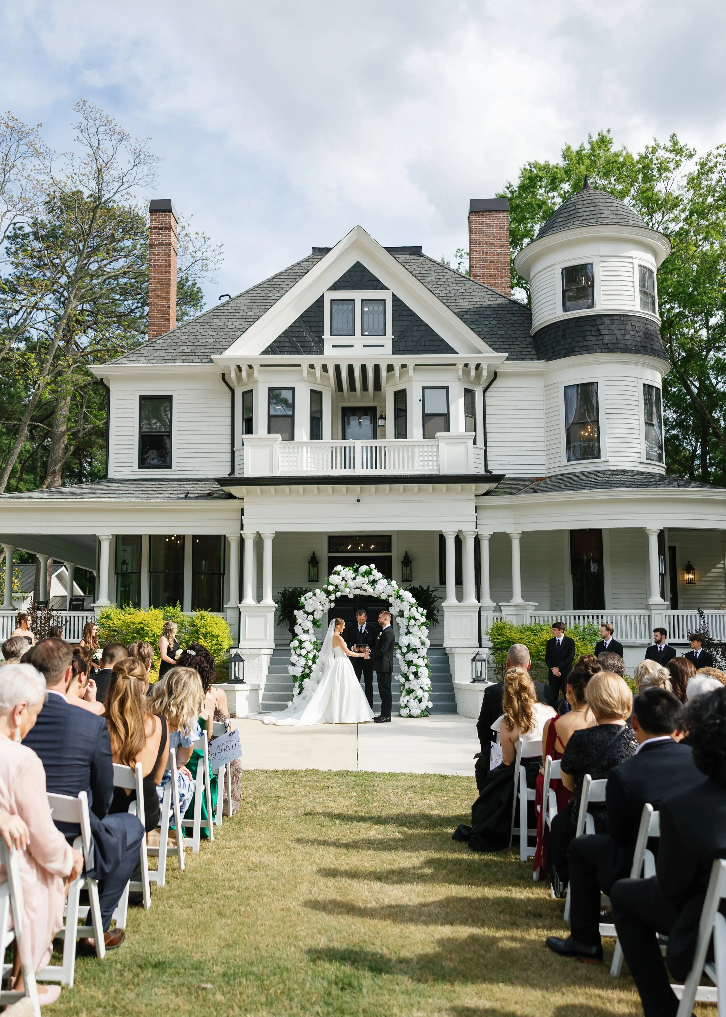 A wedding ceremony taking place outdoors in front of a large, white Victorian-style house with a turret. The bride and groom stand under a floral arch, surrounded by guests seated in rows on a grassy lawn.