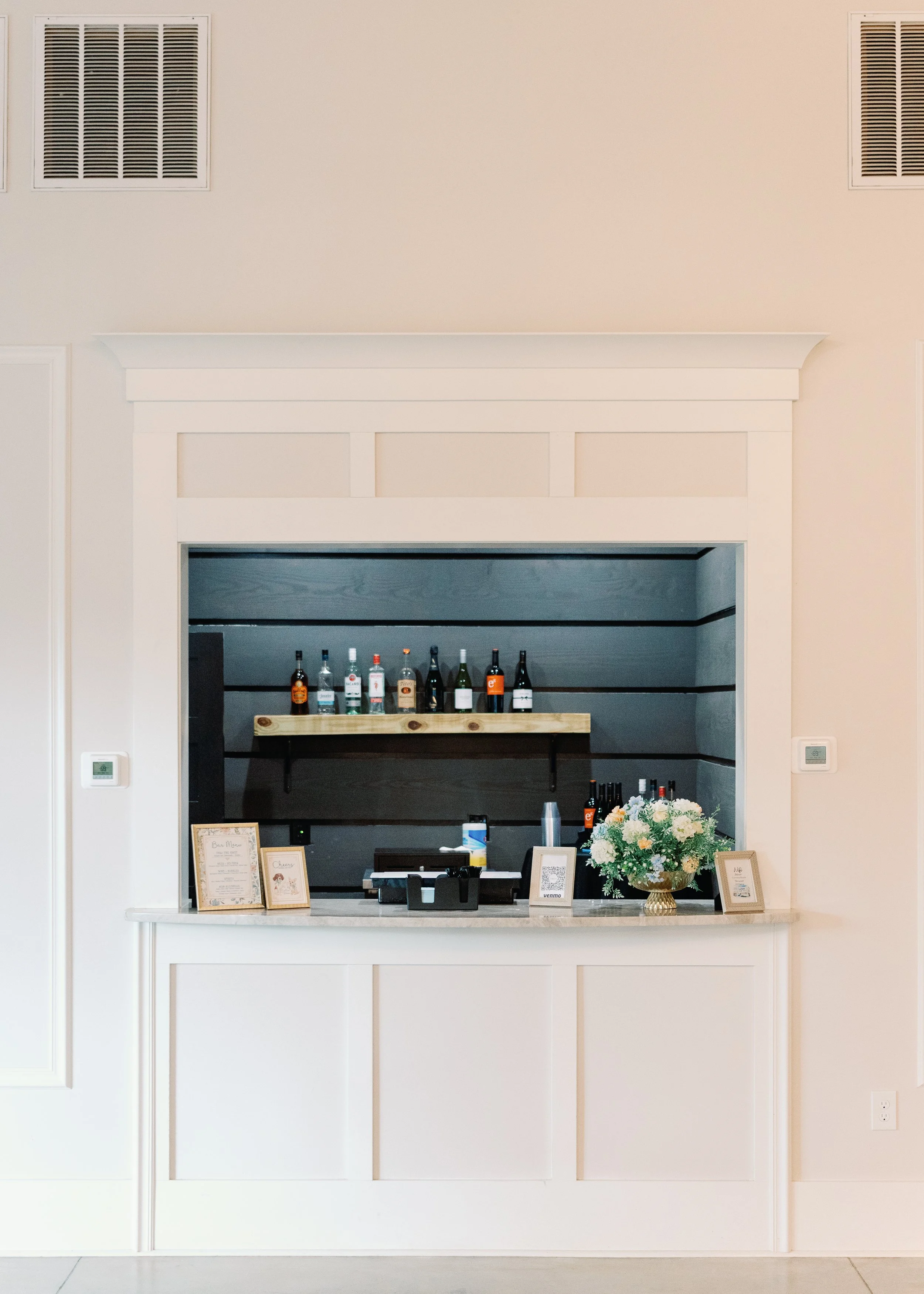 A bar section with bottles of alcohol on a wooden shelf, framed pictures, and a bouquet of flowers on a countertop.