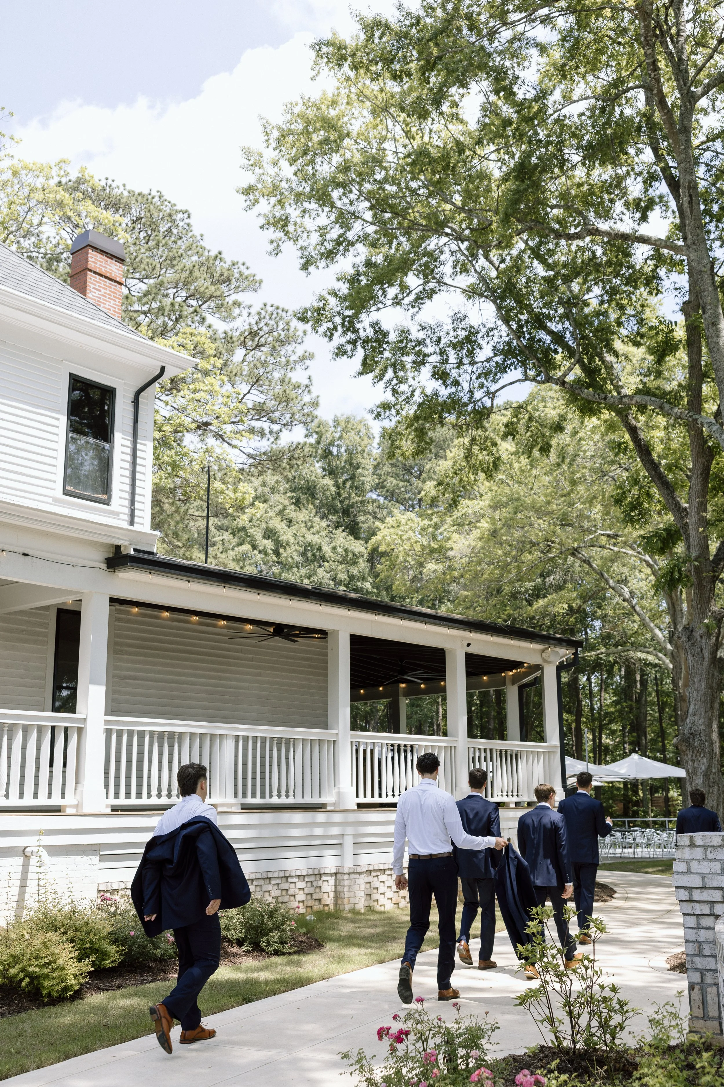 A group of six men dressed in suits walking on a sidewalk towards a white house surrounded by trees and greenery, with a covered porch and outdoor seating area.