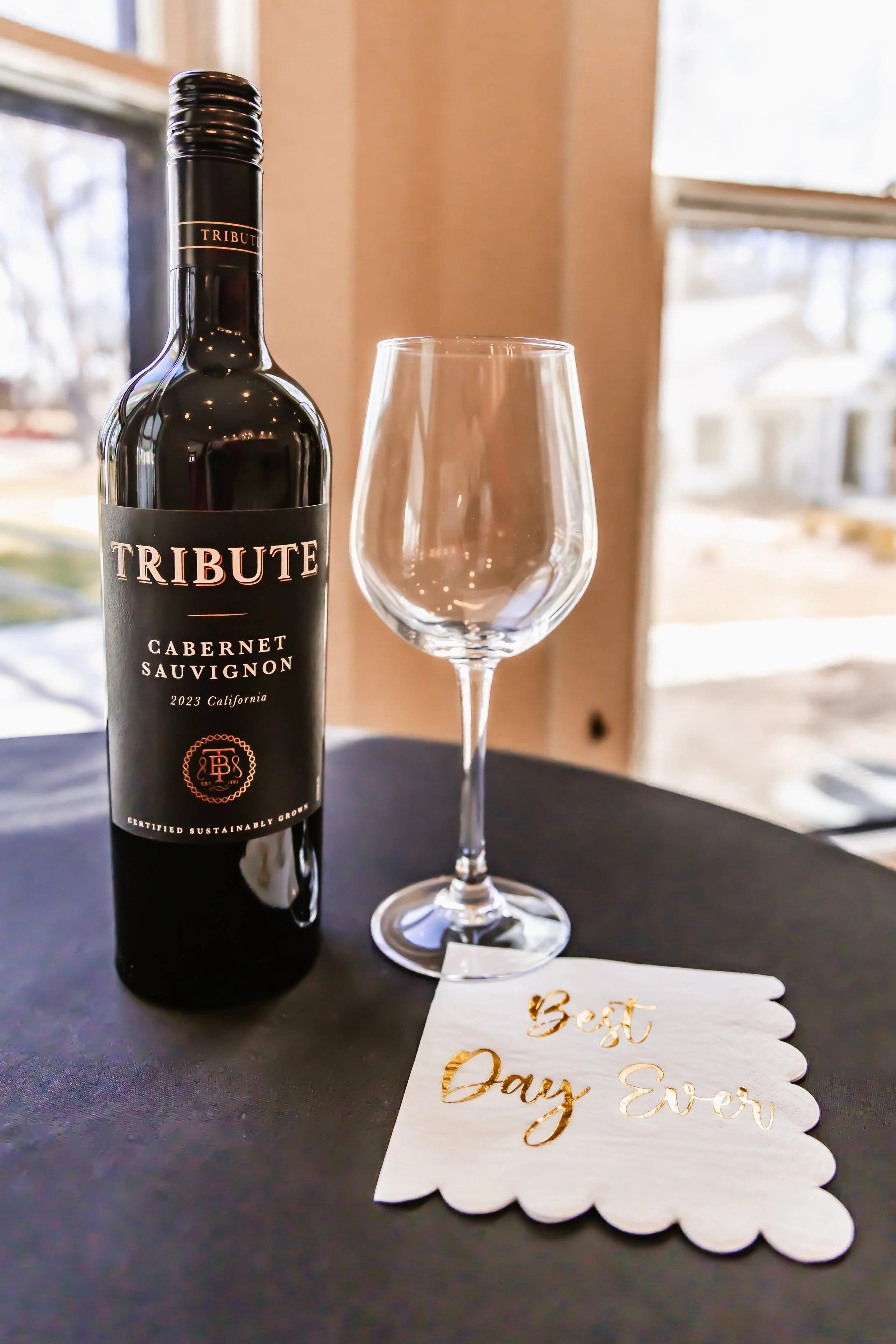 A bottle of Tribute Cabernet Sauvignon wine, an empty wine glass, and a white napkin with gold writing that says "Best Day Ever" on a black table near a window.