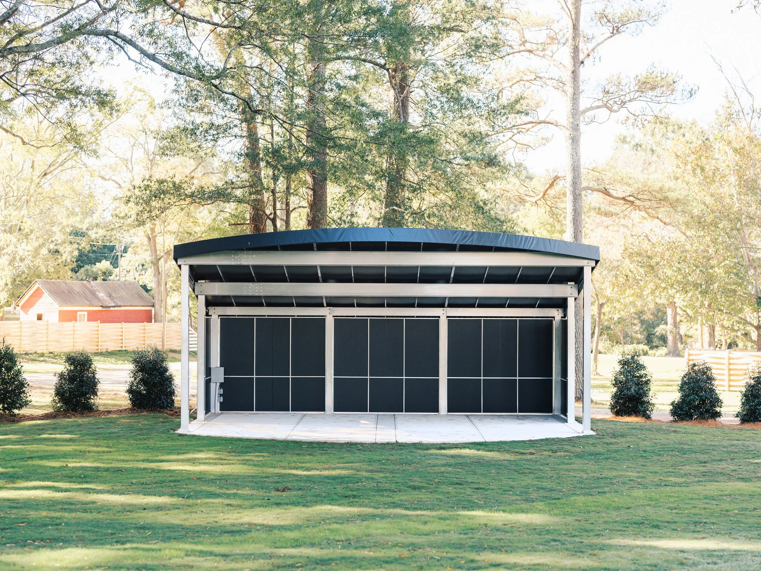 A black and gray outdoor pavilion with a curved roof in a grassy park surrounded by trees and bushes.
