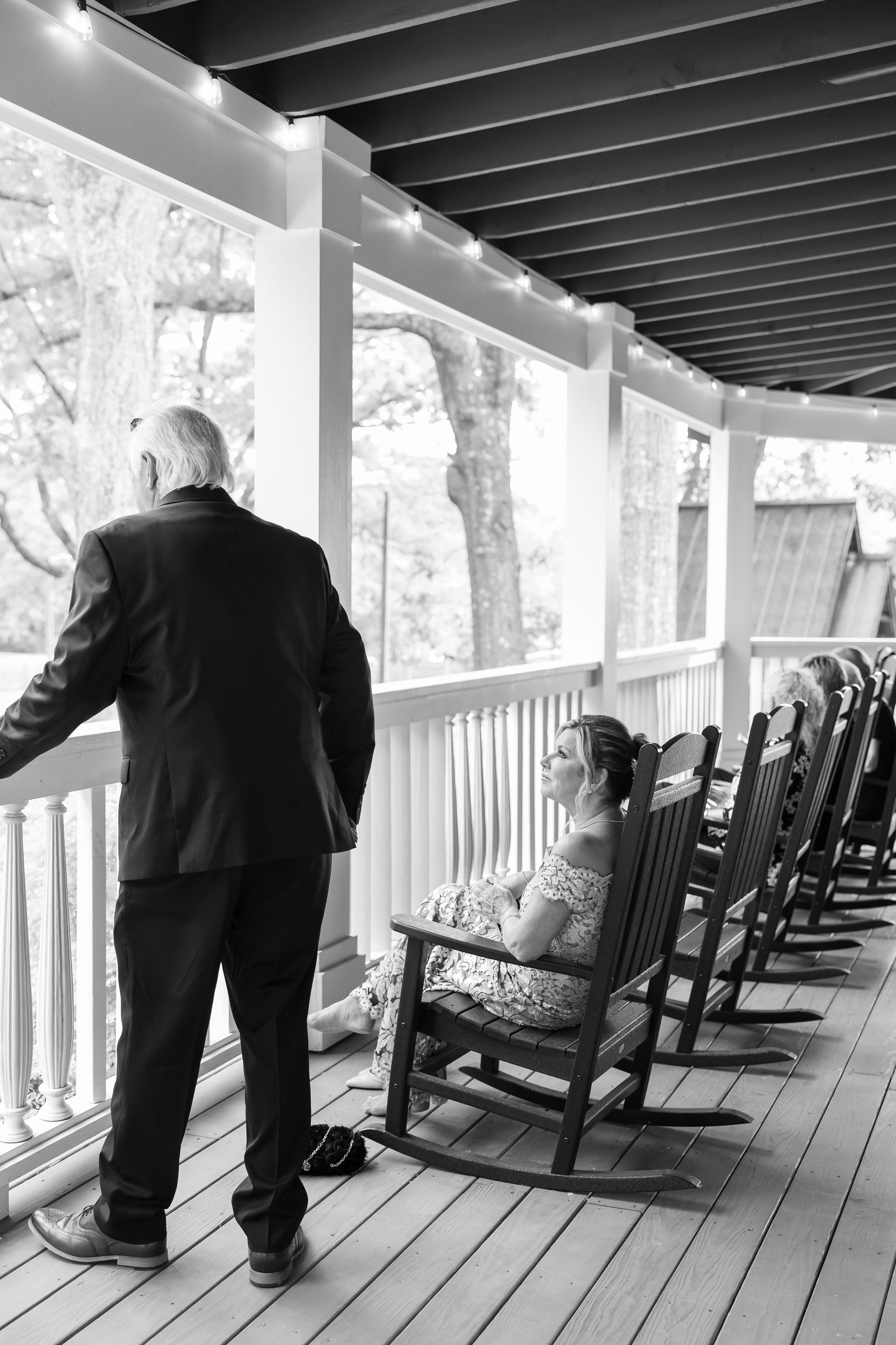 A woman in a floral off-the-shoulder dress sits in a rocking chair on a wooden porch, looking up at a man in a suit who is standing nearby, during daytime. Other chairs are lined up along the porch railing, with trees visible in the background.