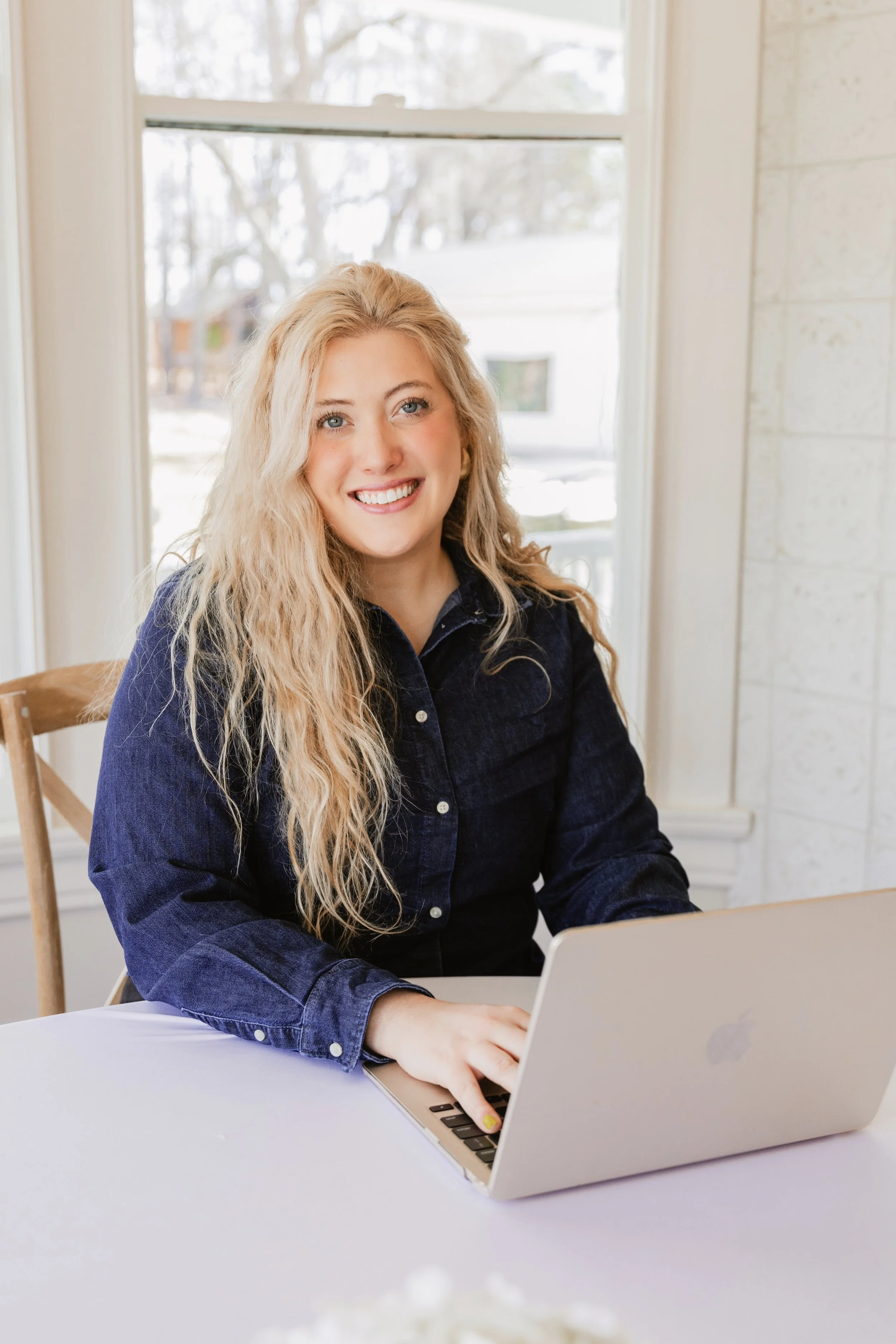 A young woman with long blonde curly hair sitting at a table, smiling at the camera with a laptop in front of her, in a well-lit room with a window and a white wall behind her.