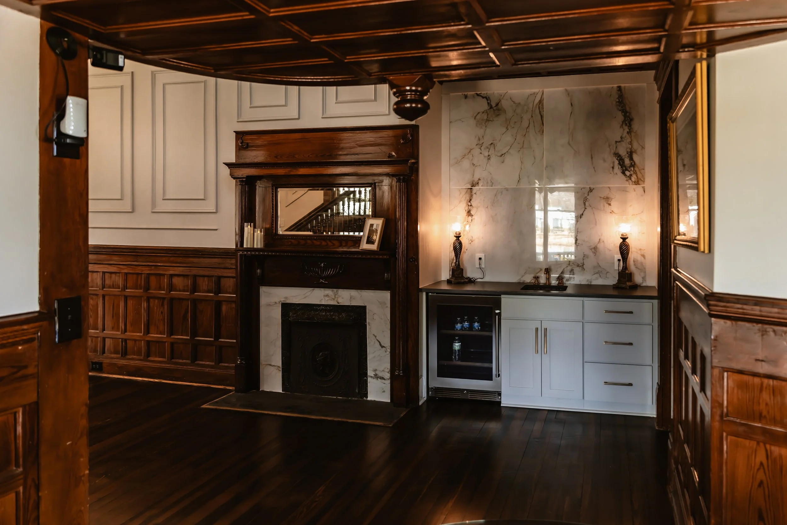 Interior of a room with a fireplace, marble wall, wood paneling, and a small white bar area with cabinets and lamps.