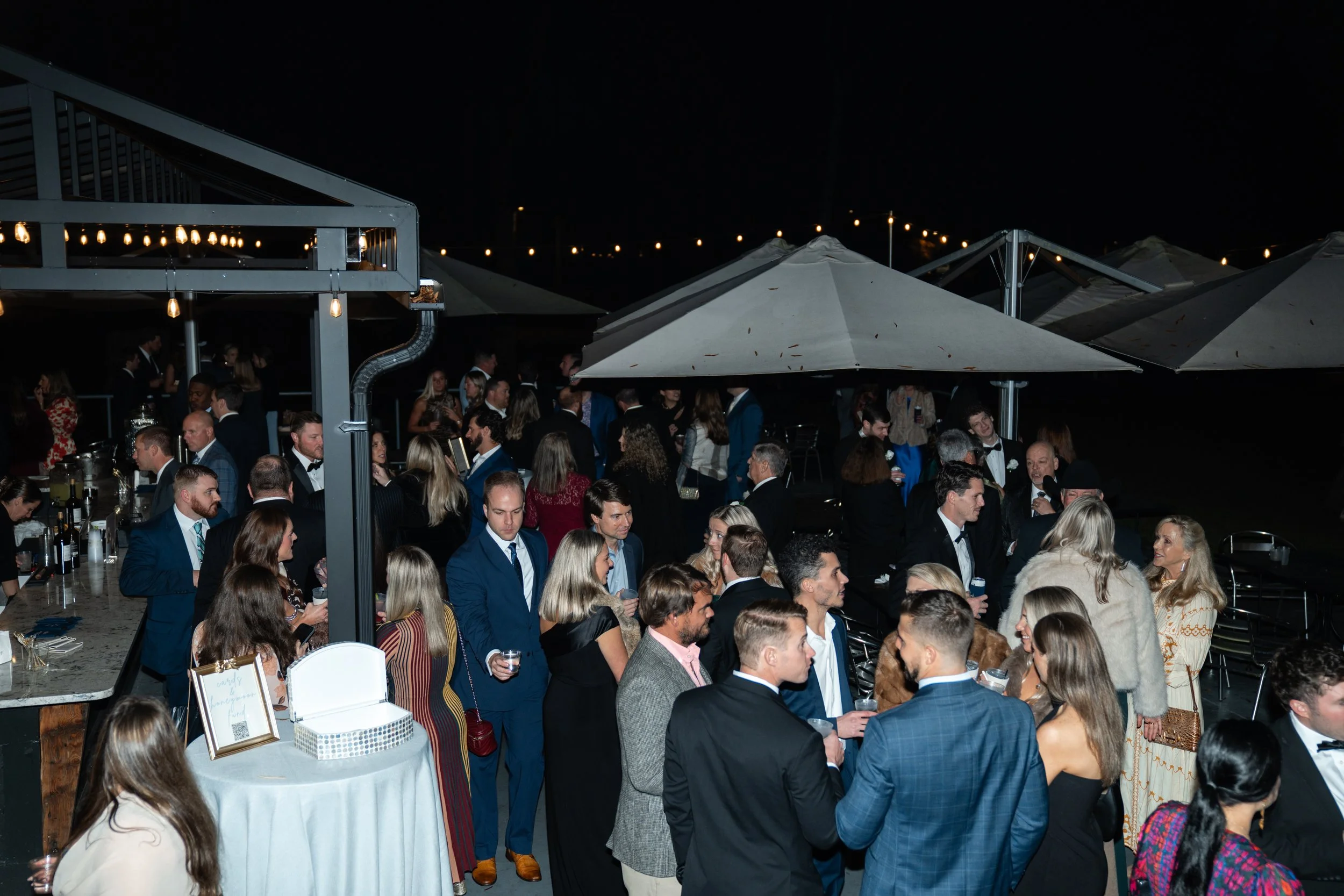 Nighttime outdoor social gathering with people dressed in formal attire under large umbrellas and string lights.
