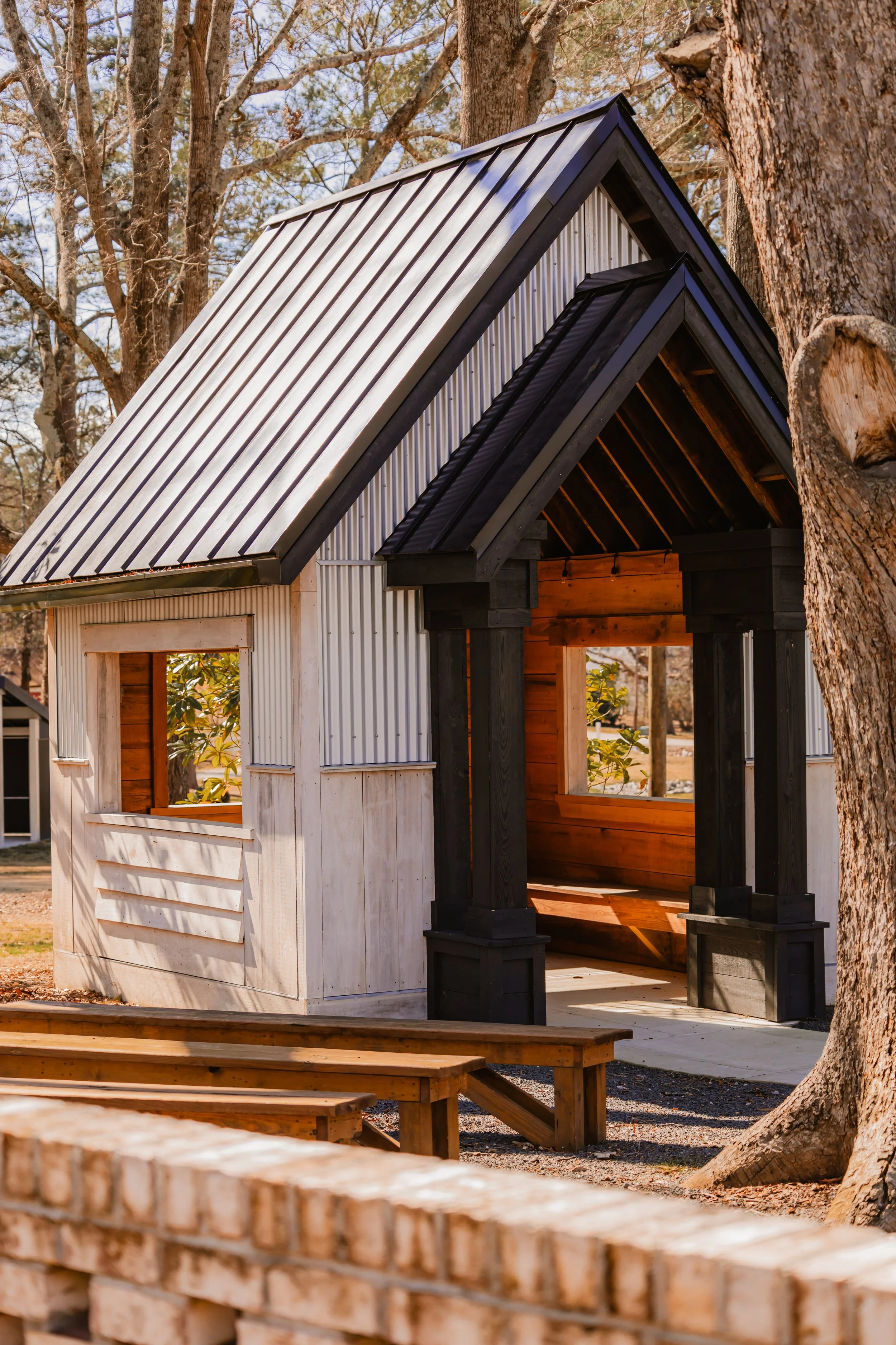 A small, partially constructed outdoor playhouse with a metal roof and wooden walls, surrounded by trees and a brick barrier.