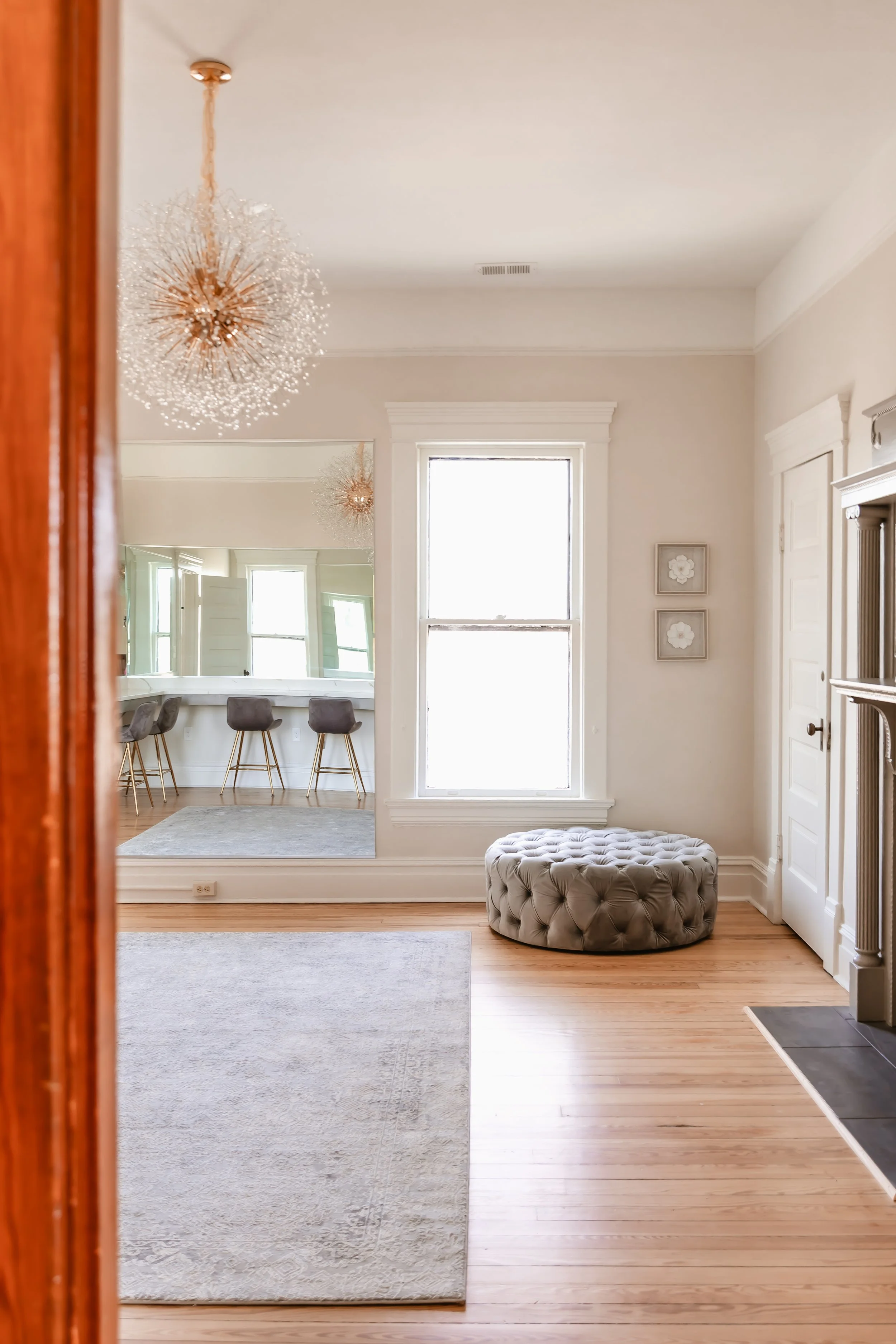 View of a bright living room with light-colored walls, hardwood floor, two large windows, a grey tufted ottoman, and a partial view of a kitchen area with chairs near a counter.