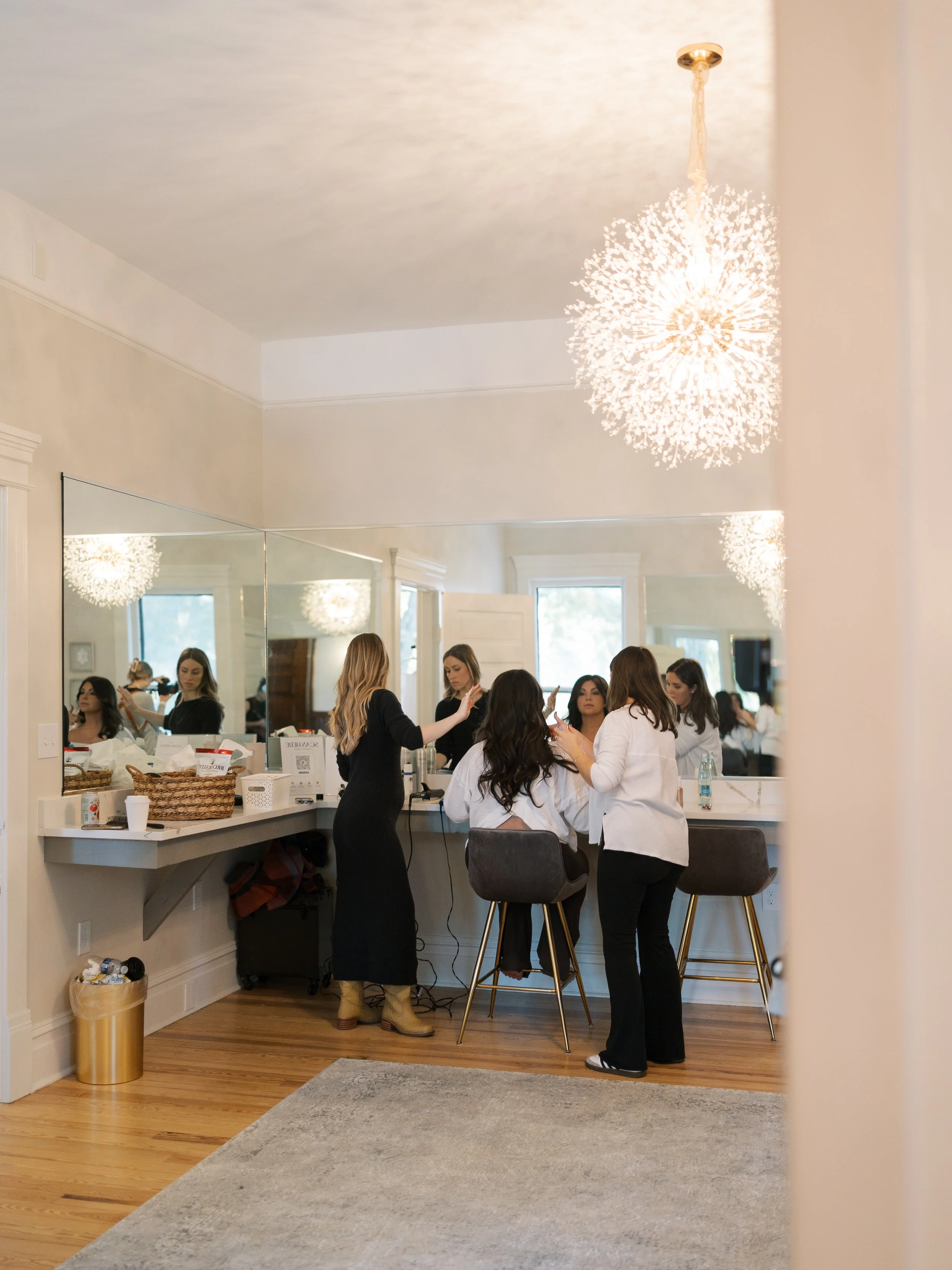 A woman getting her hair styled in front of a large mirror while another woman assists, in a well-lit room with a modern chandelier, wooden floors, and a light-colored wall.