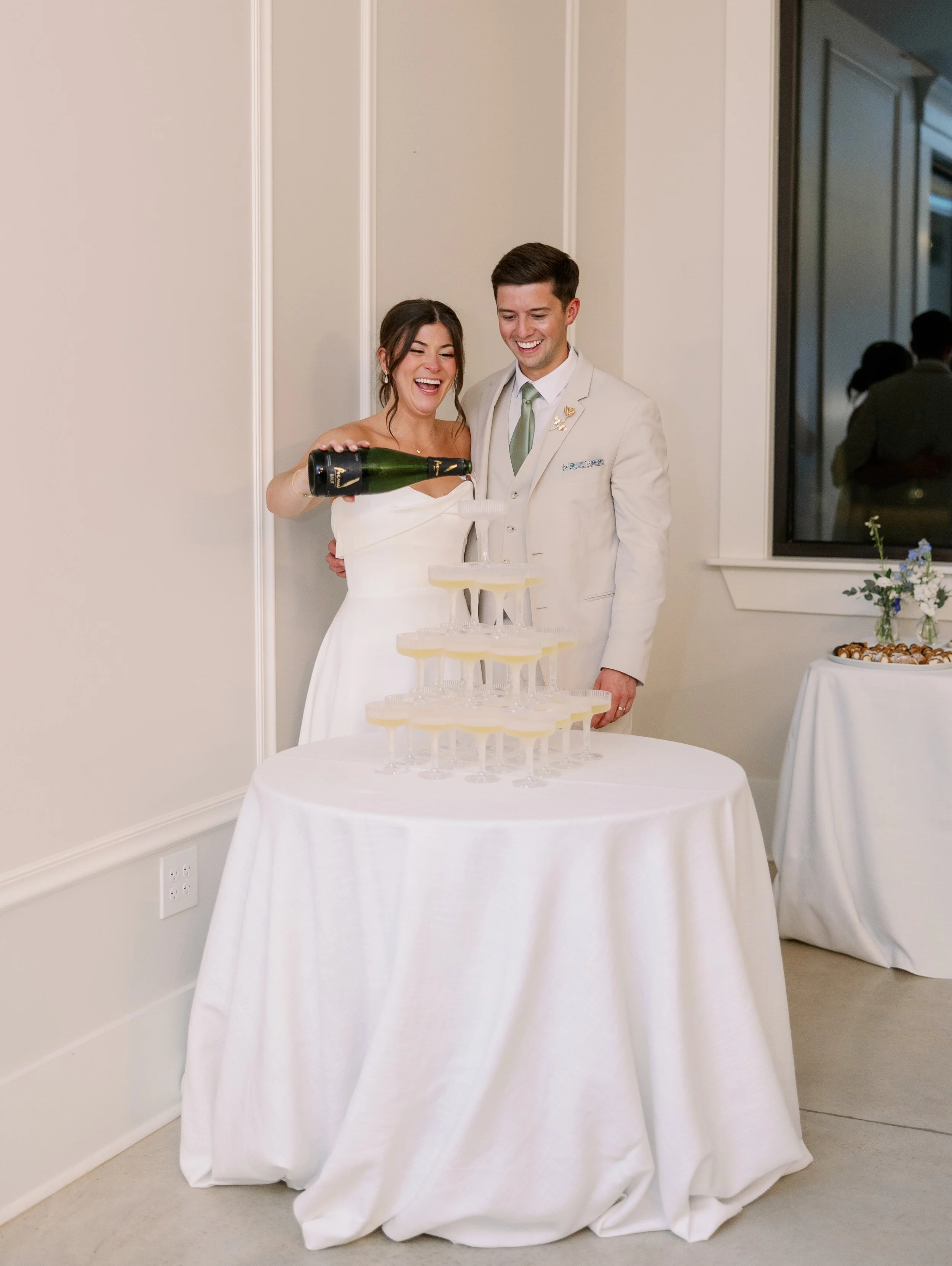 A bride and groom pouring champagne into a tower of glasses at a wedding reception, smiling and celebrating.