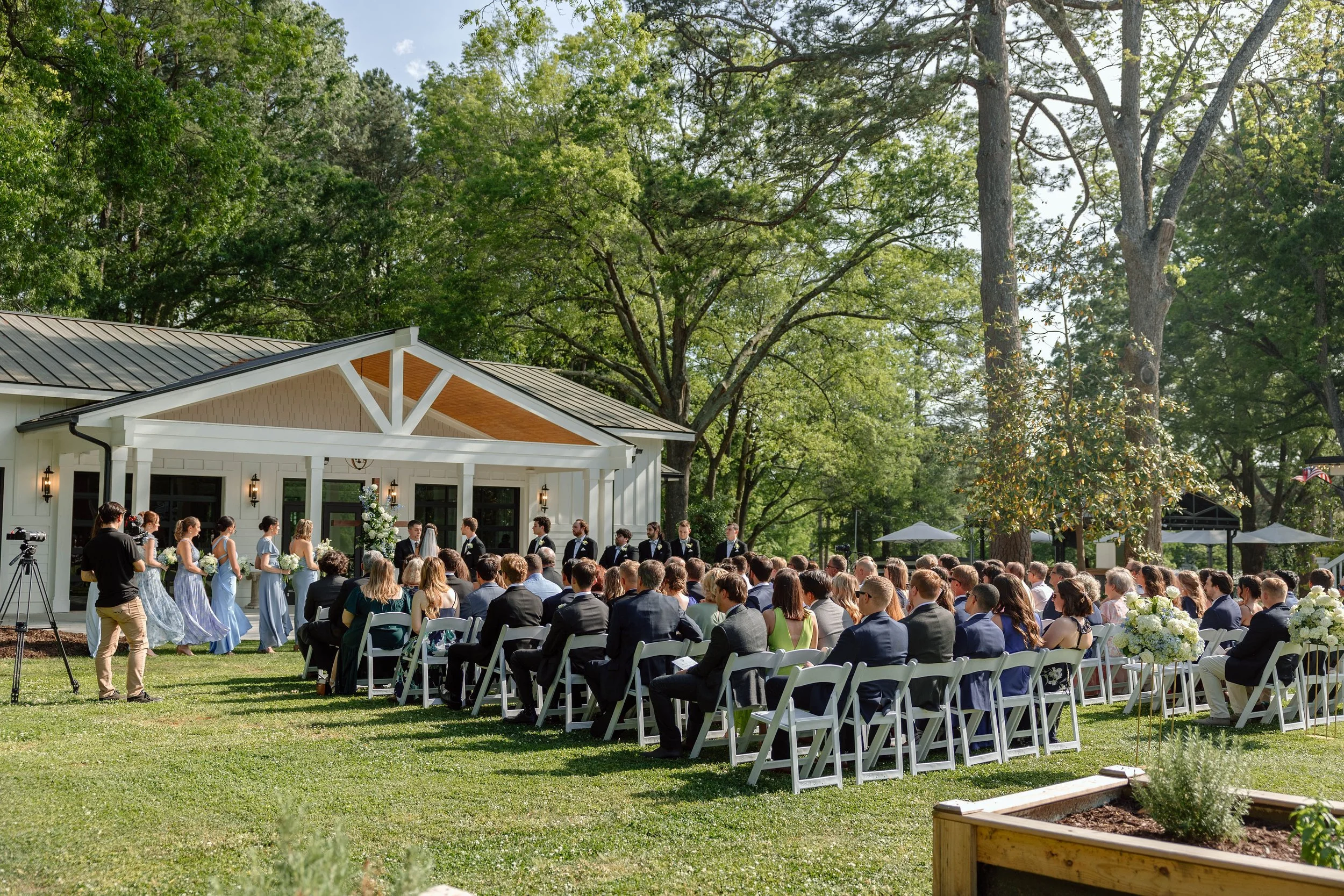 Outdoor wedding ceremony with guests seated on white chairs, bridal party standing at the front, and a backdrop of trees and a white building with porch.