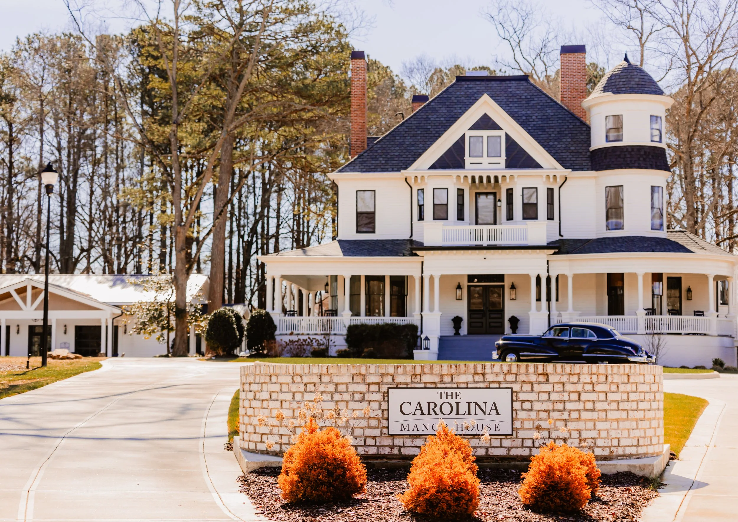 Large white mansion with dark roof, porches, and tower, surrounded by trees. A brick sign in front reads "The Carolina Mansion House" with orange bushes below.