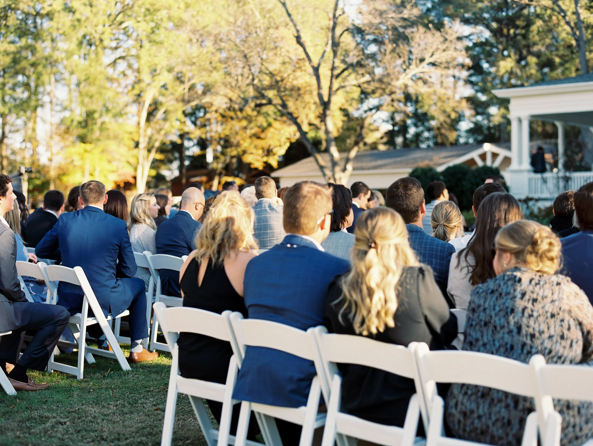 A large outdoor gathering of people seated on white chairs facing a small stage or speaker area, outdoors on a sunny day with trees and a house in the background.